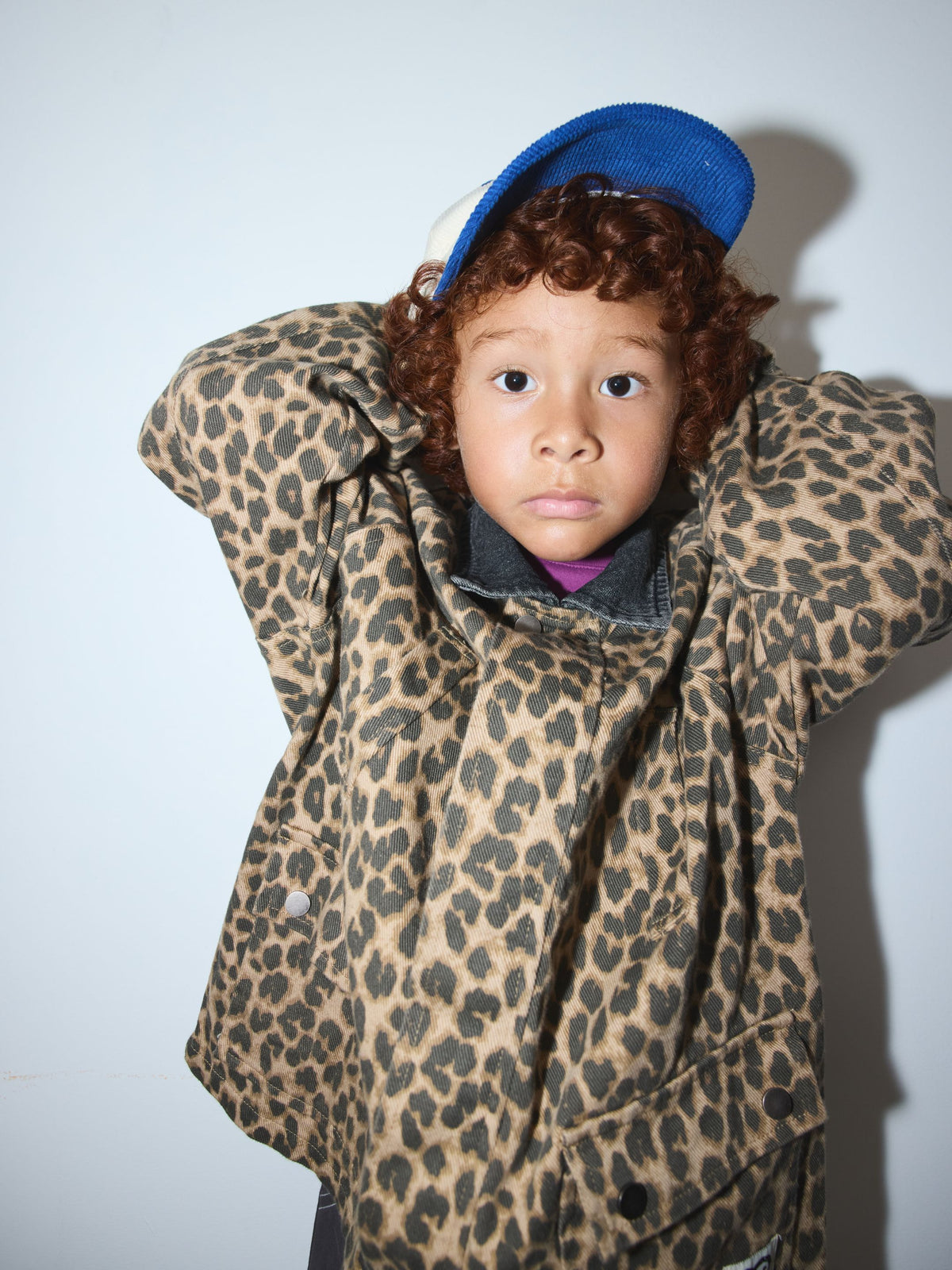 Leopard | A child wearing a leopard-print jacket and a blue cap, posing with hands behind their head against a plain light background.
