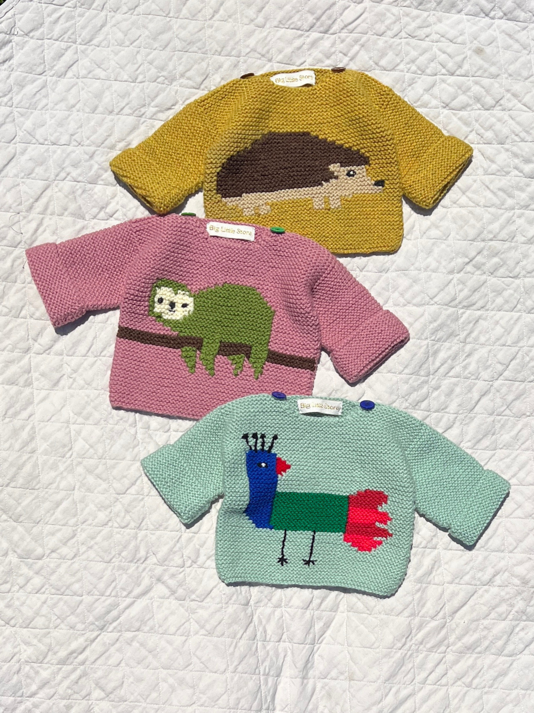 Three baby sweaters in mint green featuring a bird, pink with a sloth and ochre with a hedgehog, all laid on a white quilt background.