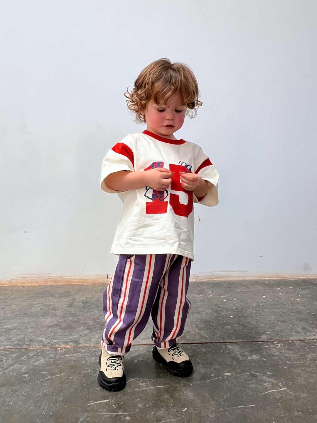 A young child with curly hair stands indoors on a concrete floor, wearing baggy fit STRIPE TWILL PANT, a white shirt with red stripes and numbers, and beige shoes, looking down at something in their hands.