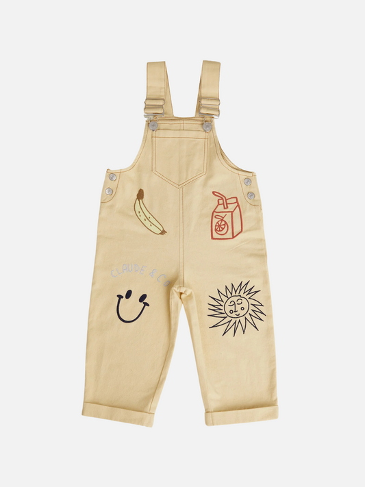 Image of The PIPER BREAKFAST DUNGAREE by Claude & Co. is a light yellow children’s overall in organic cotton twill, featuring playful banana, juice box, smiley face, and sun with a face illustrations on the front.