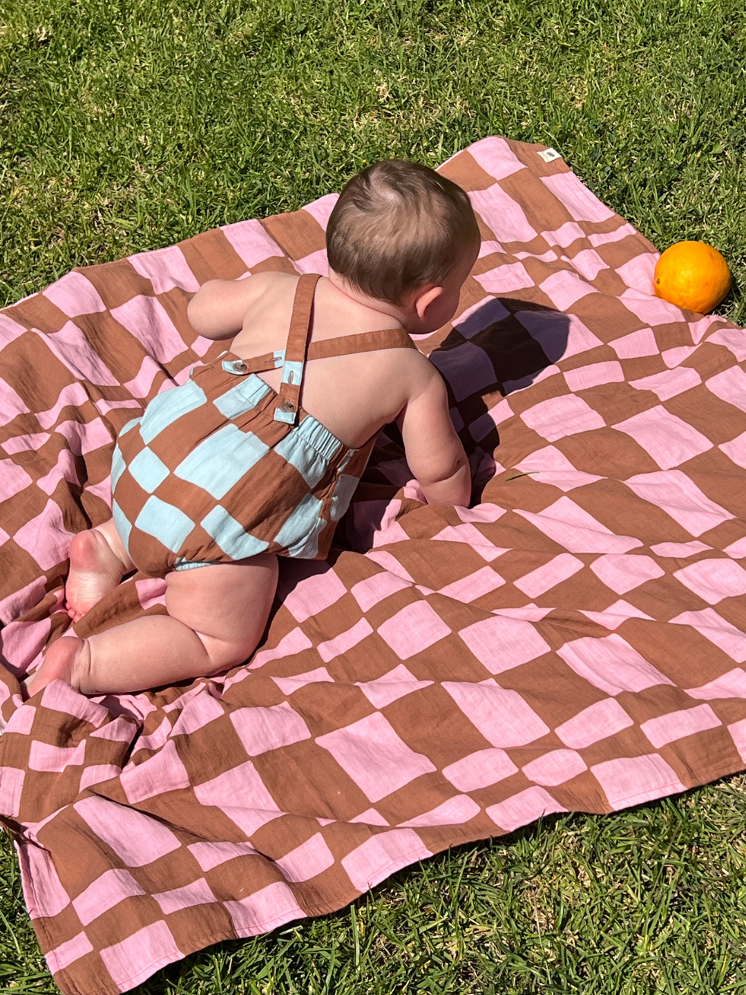 Baby in a blue and brown check romper crawling on a pink and brown checkered blanket outdoors on grass.