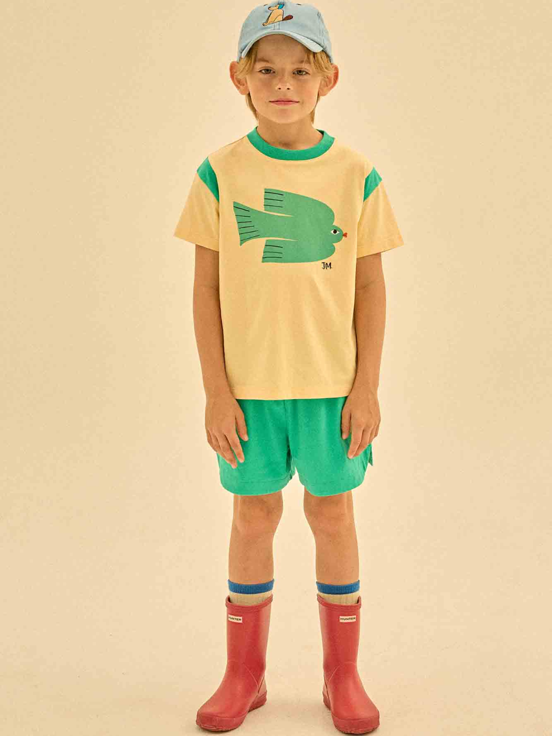 Child in a GREEN BIRD T-SHIRT, green shorts, blue cap, and red rubber boots, stands against a beige background.