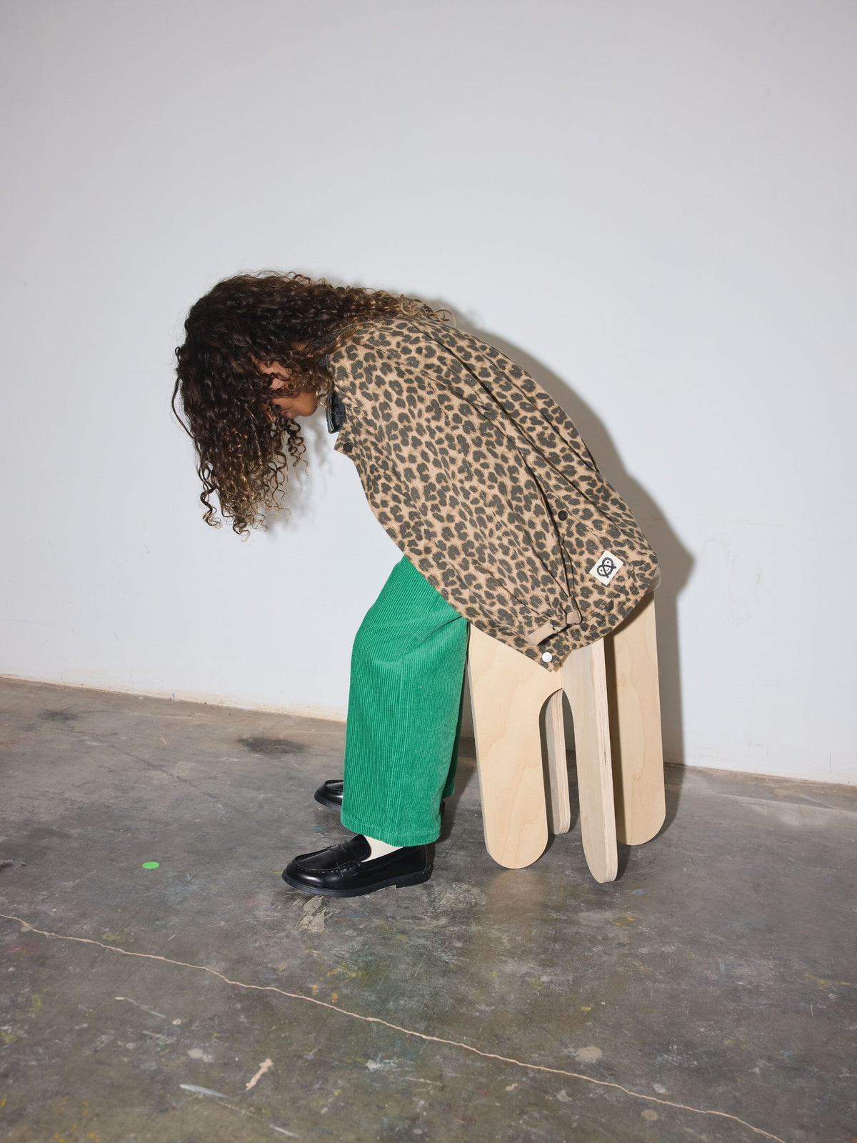A child with long curly dark hair bends forward while sitting on a light wood stool, wearing a leopard-print jacket, bright green wide-leg pants, and black loafers against a plain light gray wall and concrete floor.