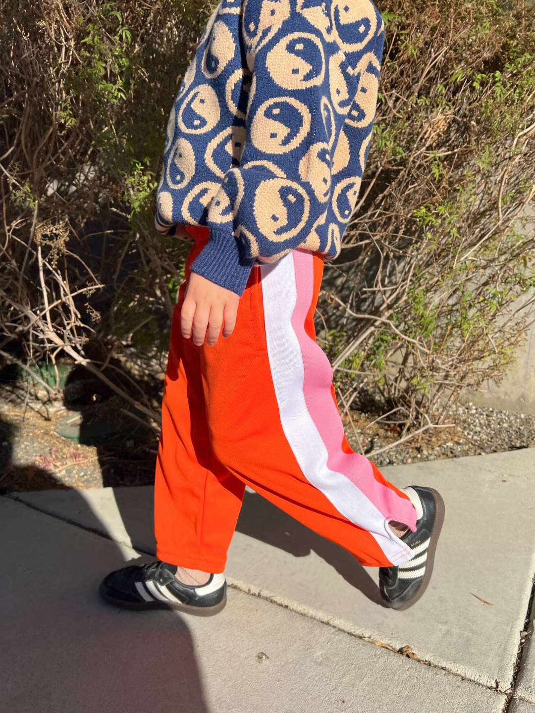 Orange | A child strolls down the sidewalk in RELAX PANTS from the Relax Lacrosse Brand, showcasing a red color with a white stripe. The ensemble is completed with a smiley face sweater and black sneakers, all set against a backdrop of lush bushes.