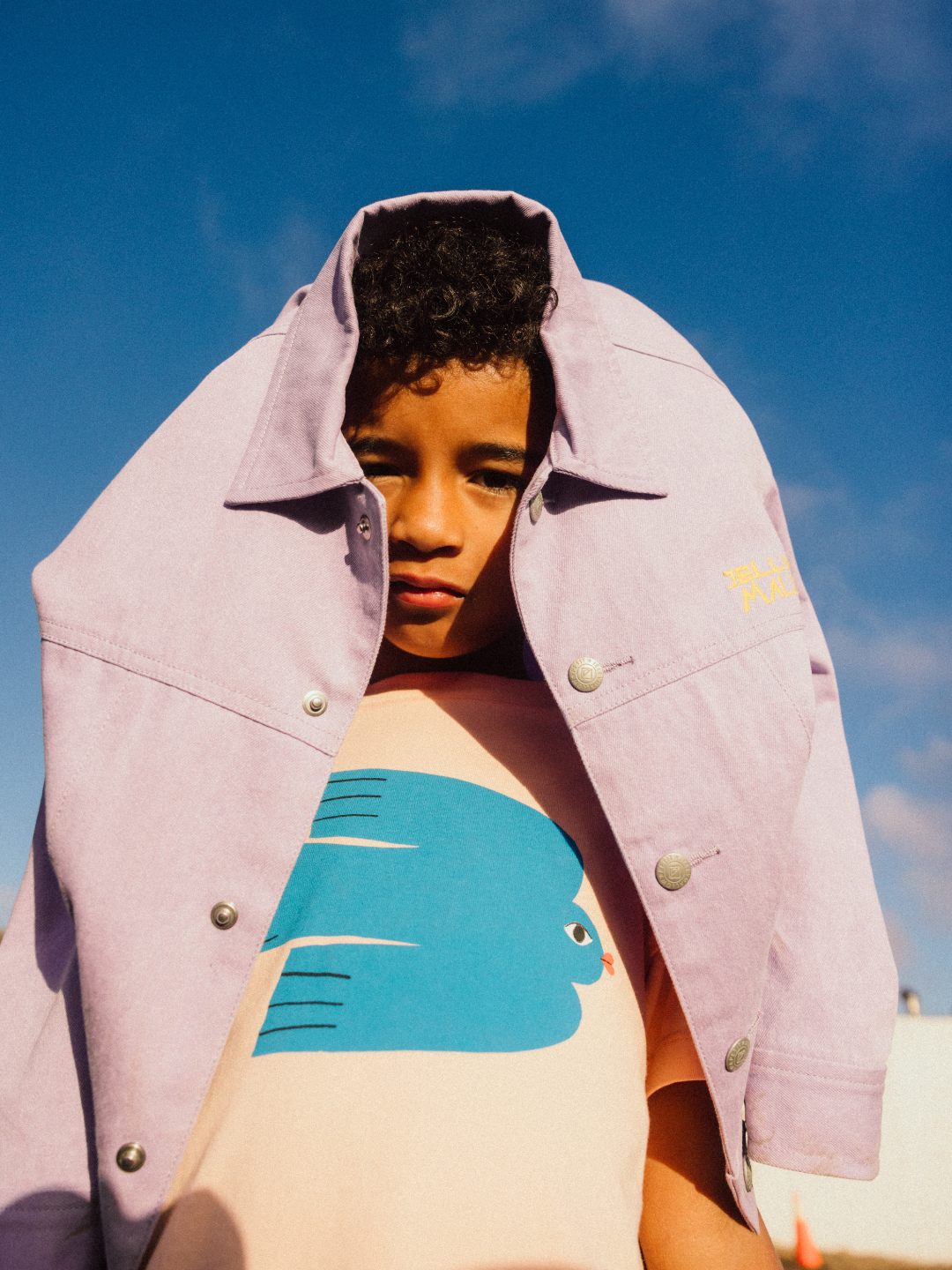 A child wearing a pink jacket draped over their head with a GREEN BIRD T-SHIRT stands against a blue sky backdrop.