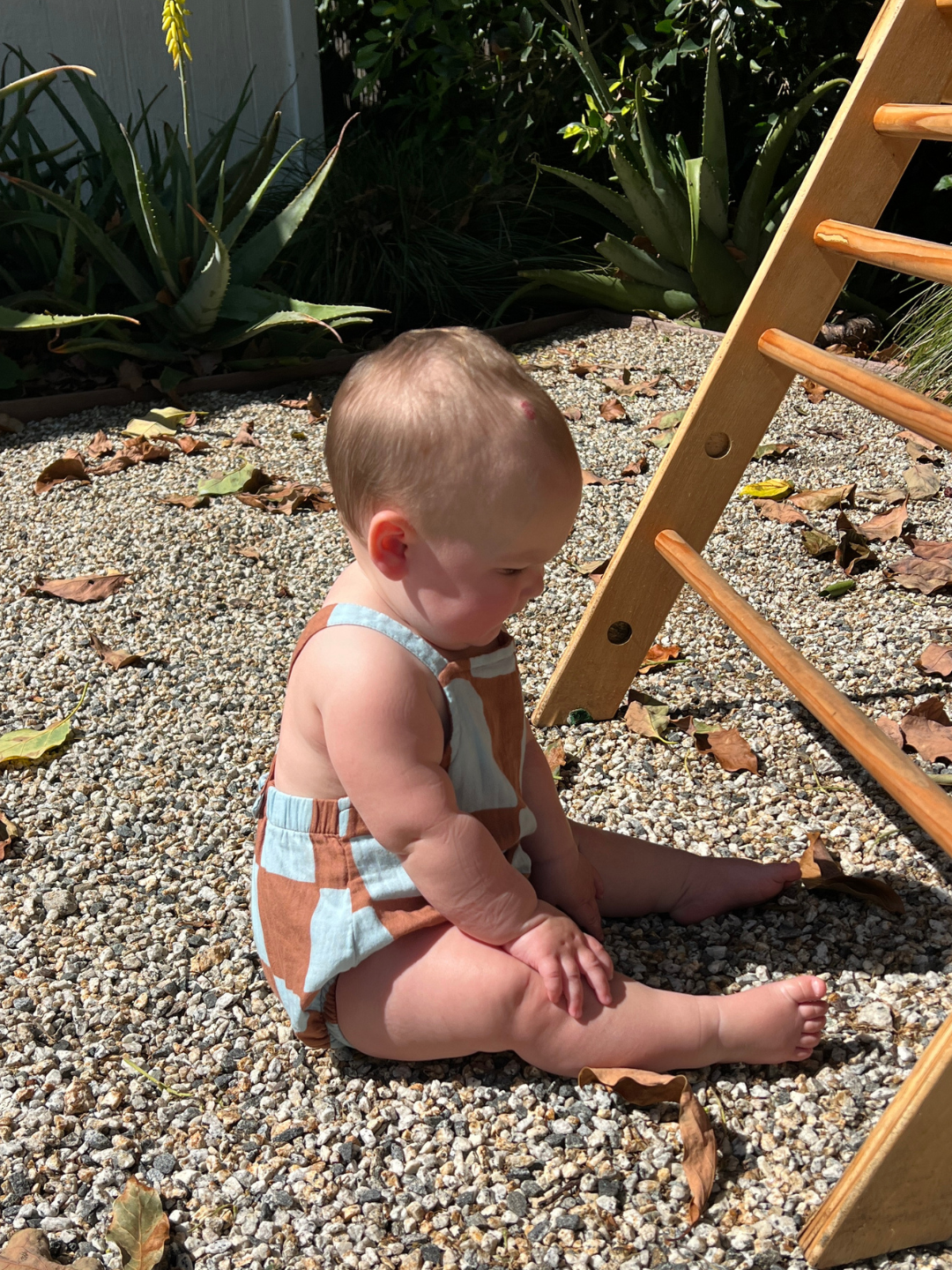 Baby in a blue and brown check romper sitting on a gravel surface with a wooden climbing toy in the background