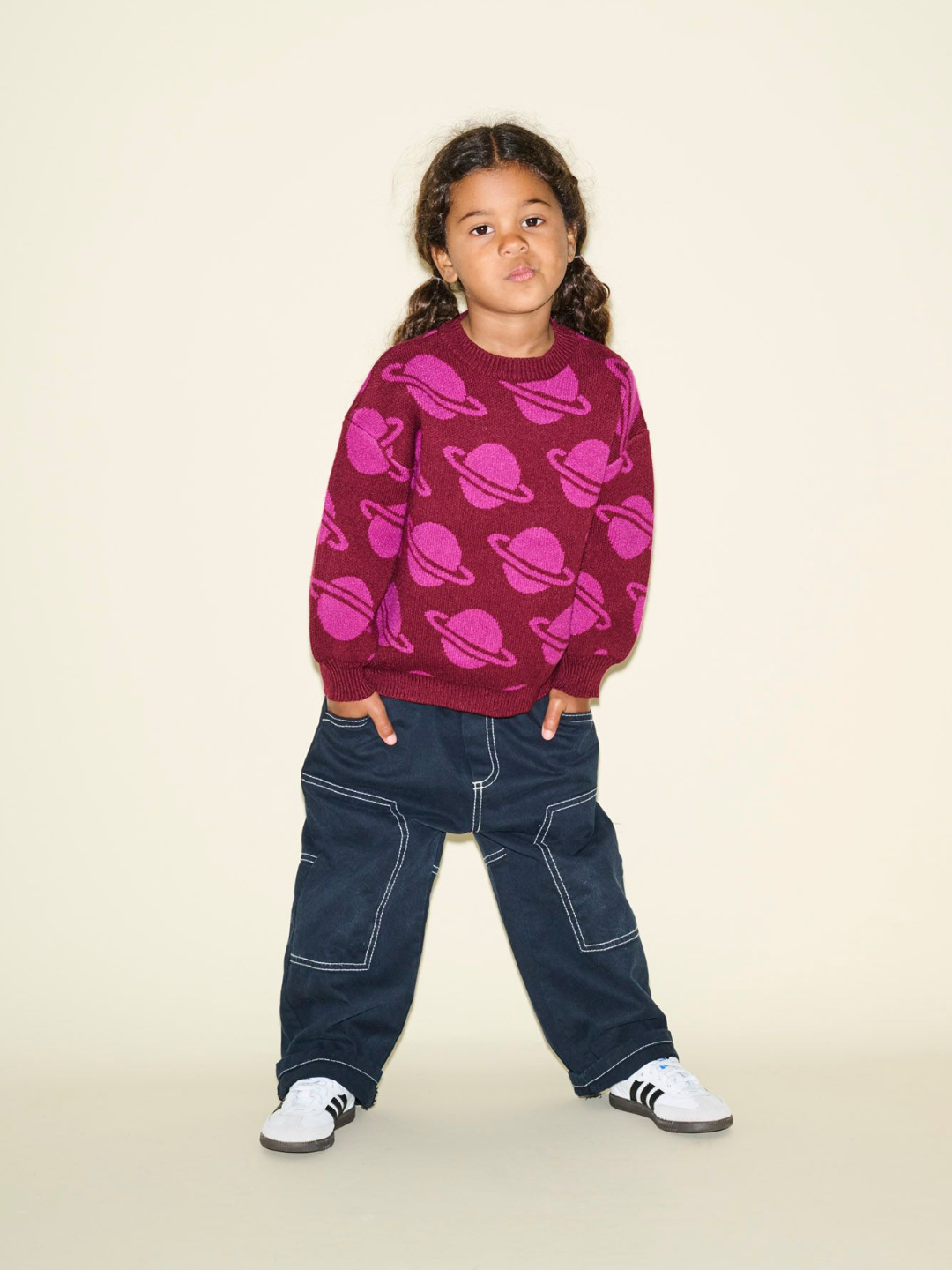 A young child with braided hair wearing a SPACE CAMP SWEATER adorned with pink planet designs, paired with dark blue jeans. The child, standing against a plain beige background with hands in pockets, seems ready for photos that capture every detail of this charming description.