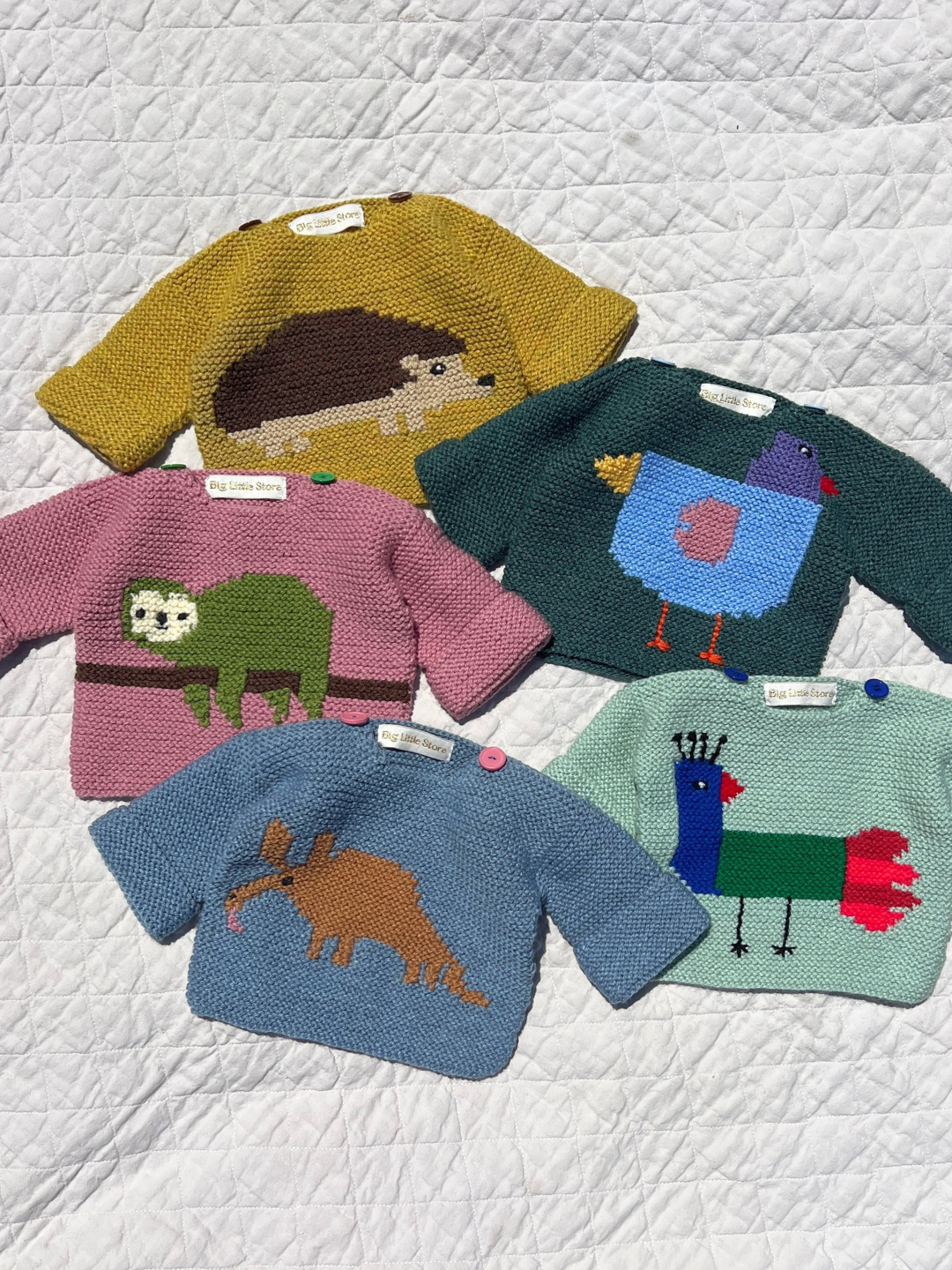 E (blue anteater) | Five baby sweaters shown flat on a white quilt background: ochre with hedgehog, teal with bird, pink with sloth, mint with bird and blue with anteater.