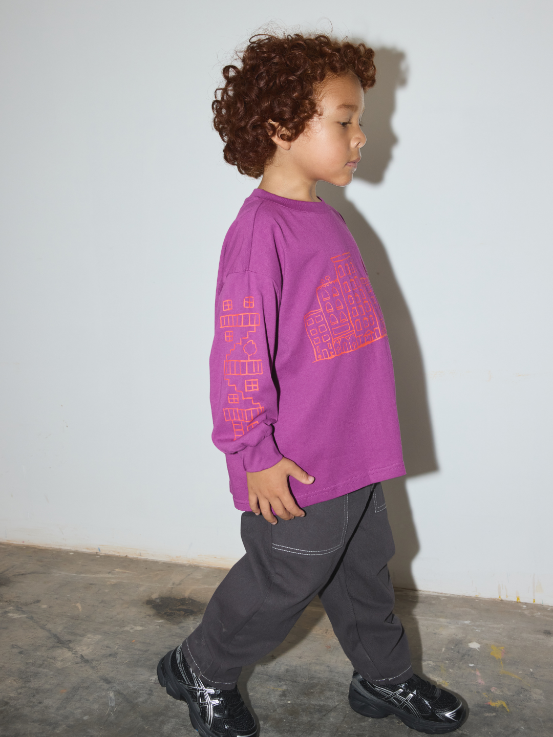 A toddler with curly brown hair is shown in profile walking, wearing a purple long-sleeve graphic t-shirt, dark gray pants, and black shoes against a plain light gray wall.