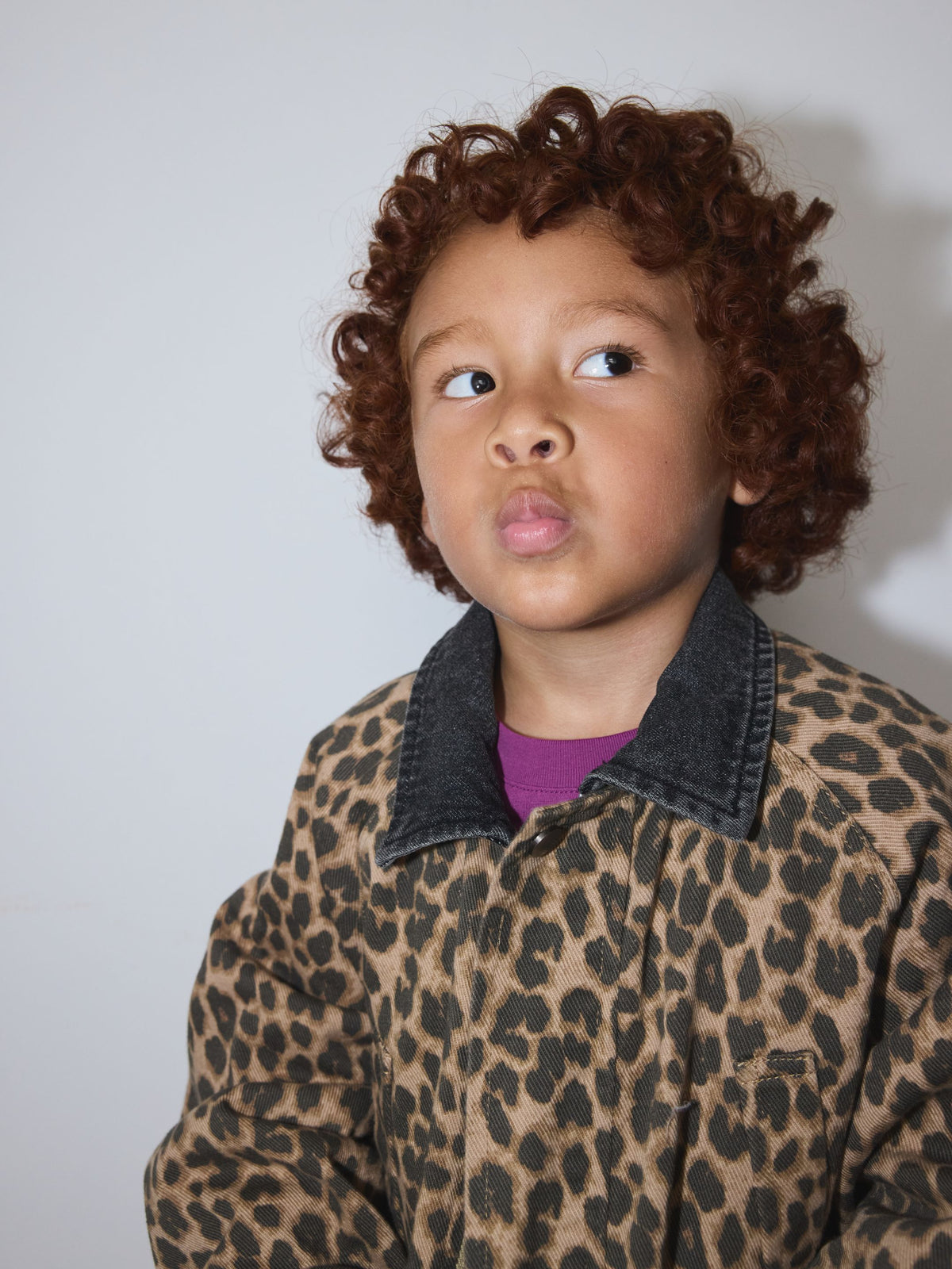 Leopard | A child with curly brown hair wearing a leopard print jacket with a dark gray collar over a purple shirt, photographed against a plain light gray background.