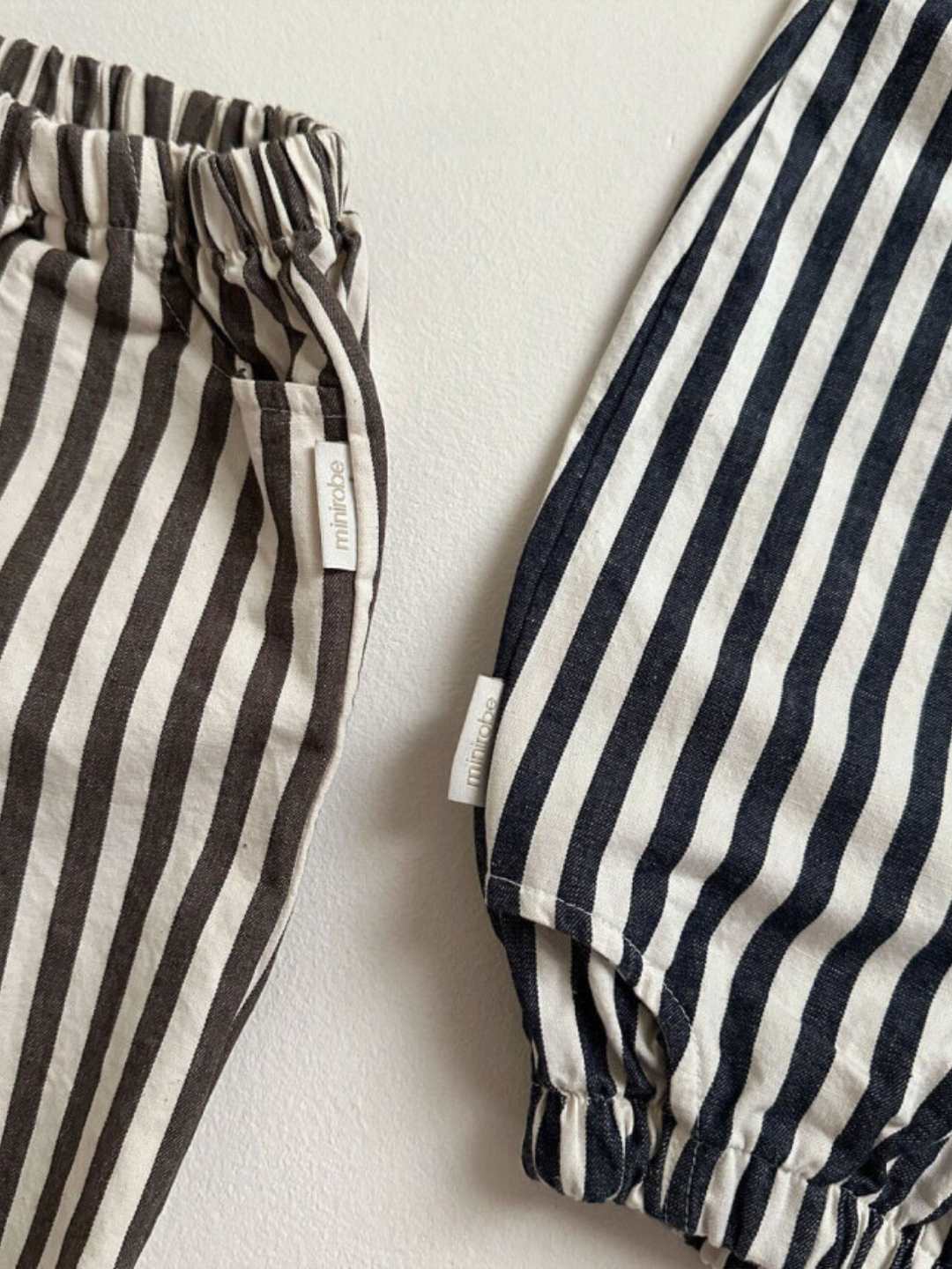 Charcoal Stripe | Close-up of two pairs of PIRATE PANTS on a surface, one featuring vertical black and white stripes and the other sporting vertical brown and white stripes. Both pants have elastic waistbands, small tags, and balloon legs from Minirobe.