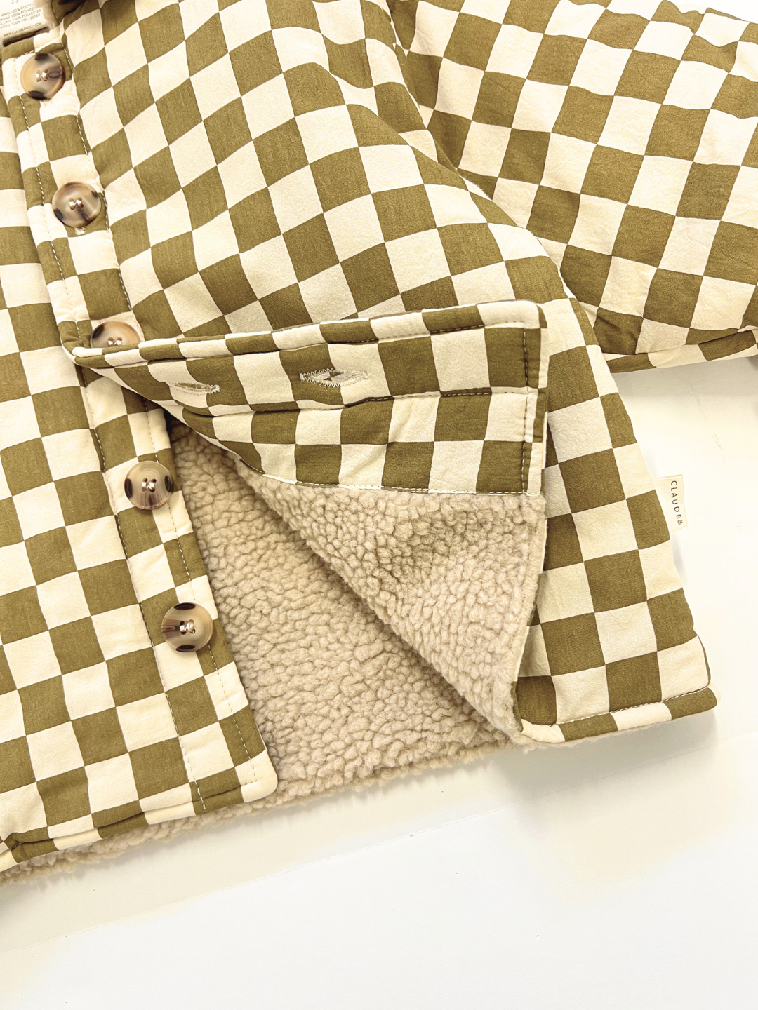 Close-up of the EMERY COCO CHECKERBOARD COAT, a unisex jacket with button closure and soft beige sherpa lining. Crafted from cotton, this oversized coat blends style and comfort—ideal for layering.