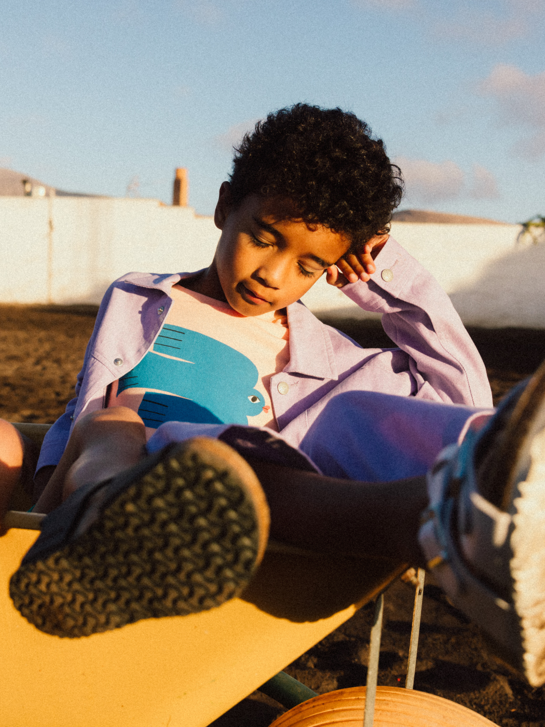 A child wearing a GREEN BIRD T-SHIRT sits casually in a wheelbarrow outdoors, leaning back with eyes closed, hand on chin, and feet extended.