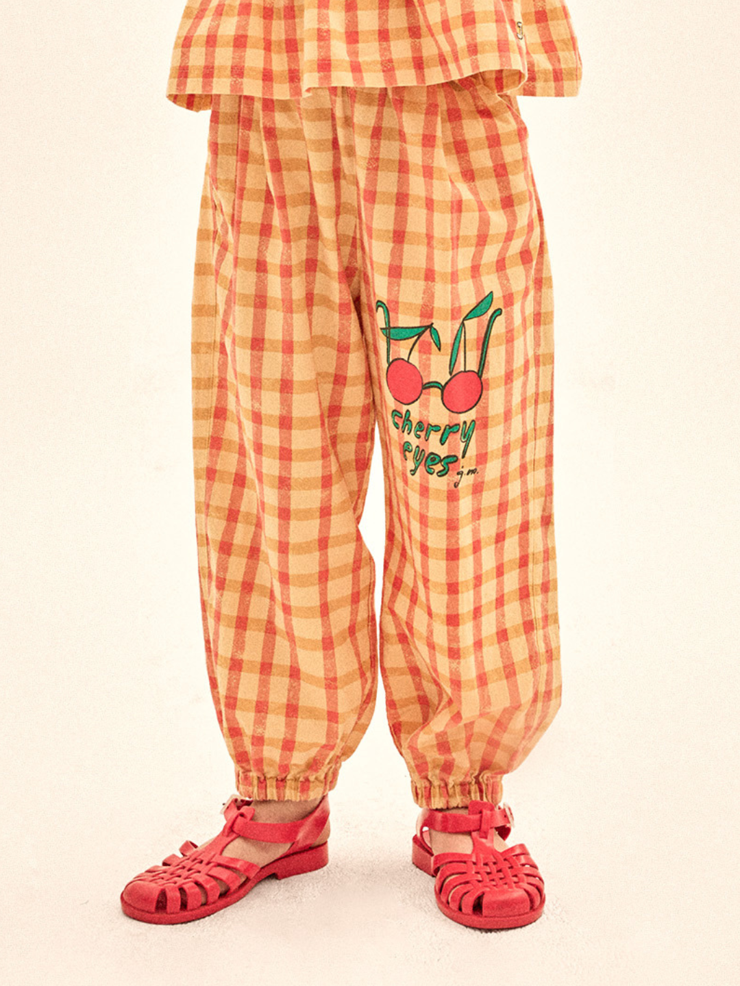 Child wearing orange checkered pants with cherry design and red sandals on a beige background