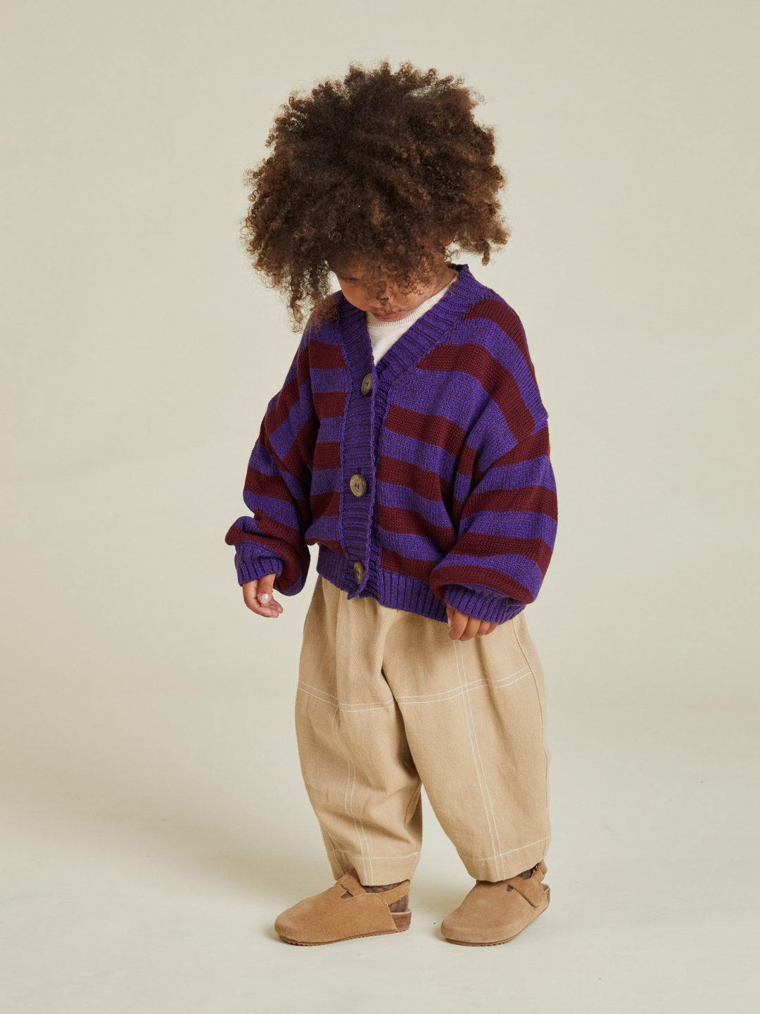 A young child wears the EBEN PURPLE STRIPE CARDIGAN with beige pants and brown shoes, standing against a plain background—perfect for cool days, plus you won’t have to worry about extra shipping cost.