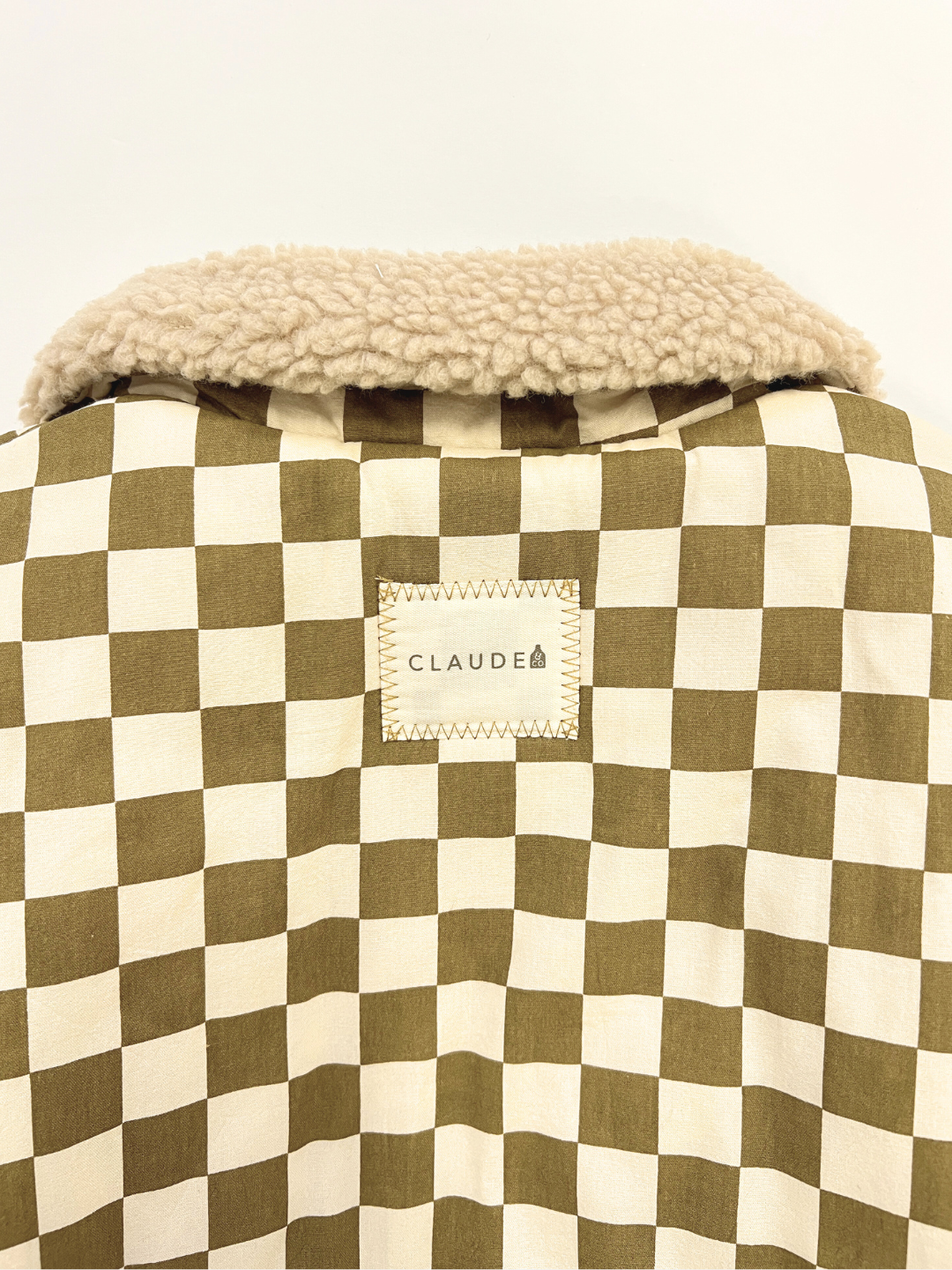 A close-up of the EMERY COCO CHECKERBOARD COAT, a unisex beige and olive cotton outerwear with a shearling collar and "CLAUDE" label—perfect for a stylish look.