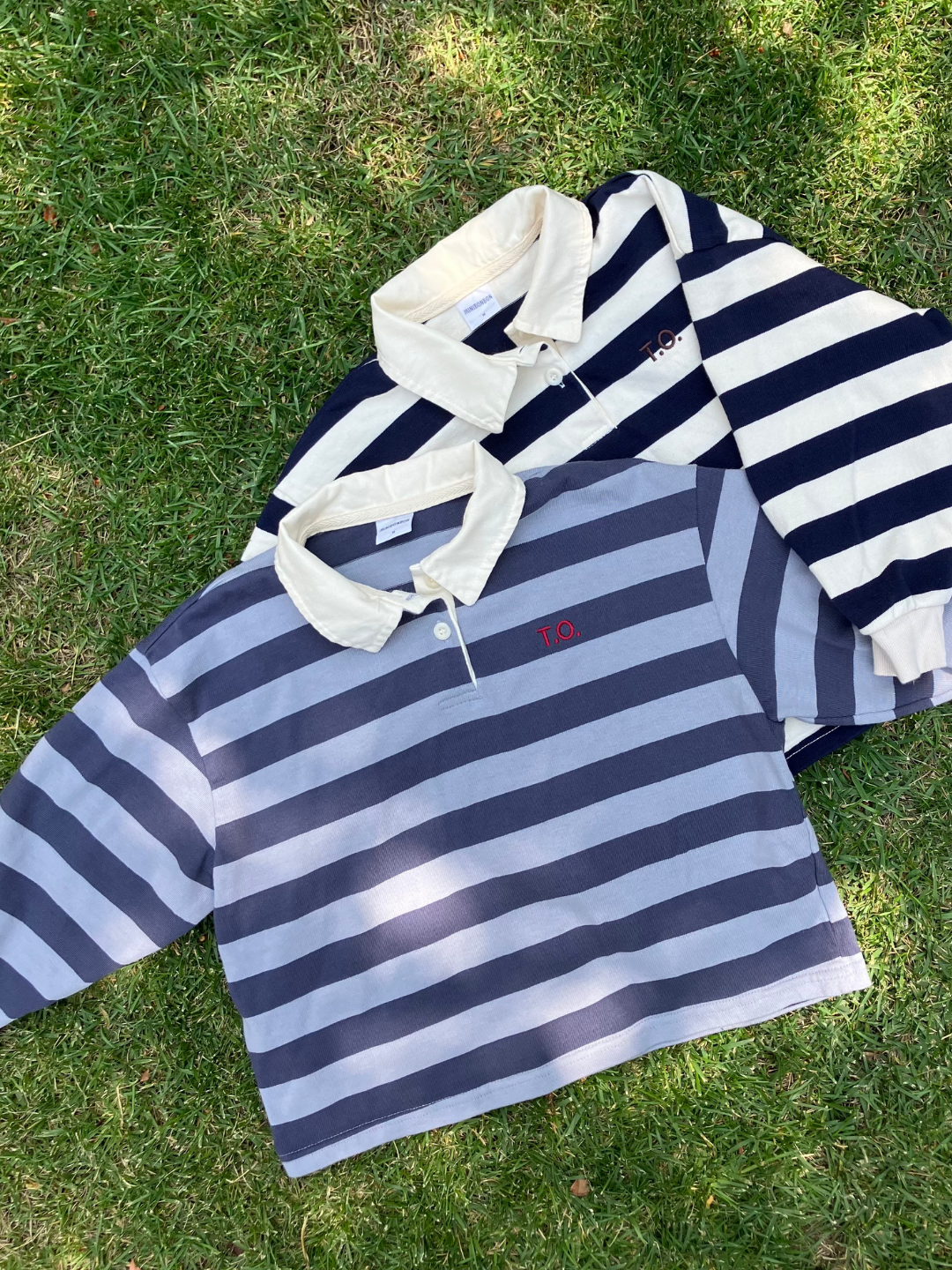 Blue | Two RUGBY LONGSLEEVE shirts made of cotton jersey fabric with horizontal stripes and collars lay on the grass. One shirt features dark blue and white stripes, while the other has light blue and dark blue stripes. Both have embroidered logos on the left chest, highlighting the quality of a popular Korean kids' brand.