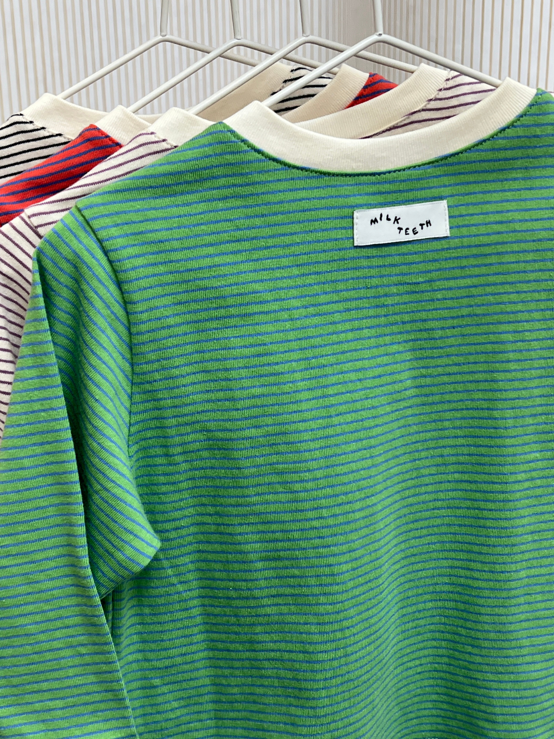 Green/Blue | Three BABY STRIPE LONGSLEEVE shirts hang on white hangers; the front one is green cotton jersey labeled "Milk Teeth," while the others feature red and black stripes—perfect for babies and toddlers, designed for easy dressing.