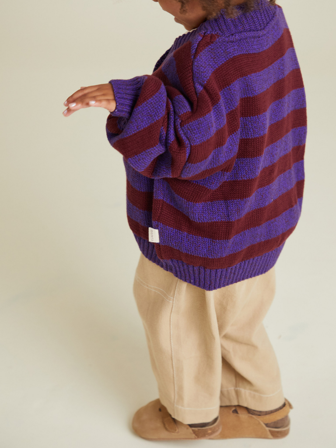 A child wears the EBEN PURPLE STRIPE CARDIGAN with tan pants and brown shoes, standing against a plain background. No additional shipping cost or tariffs apply.