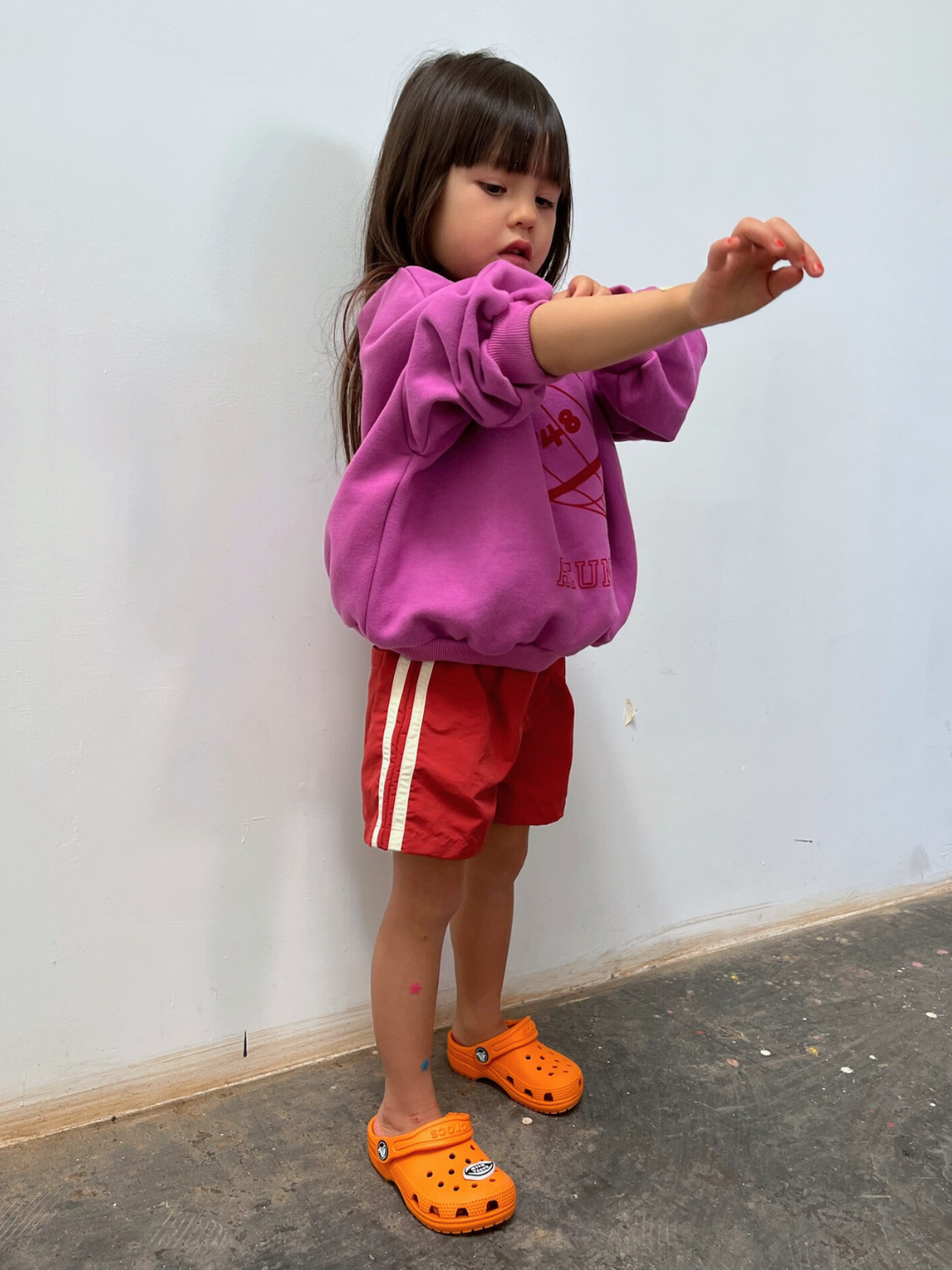 Vintage Red | A young child wearing a purple sweatshirt, SPRINTER SHORTS with white stripes, and orange clogs stands indoors against a plain wall, adjusting their sleeve.