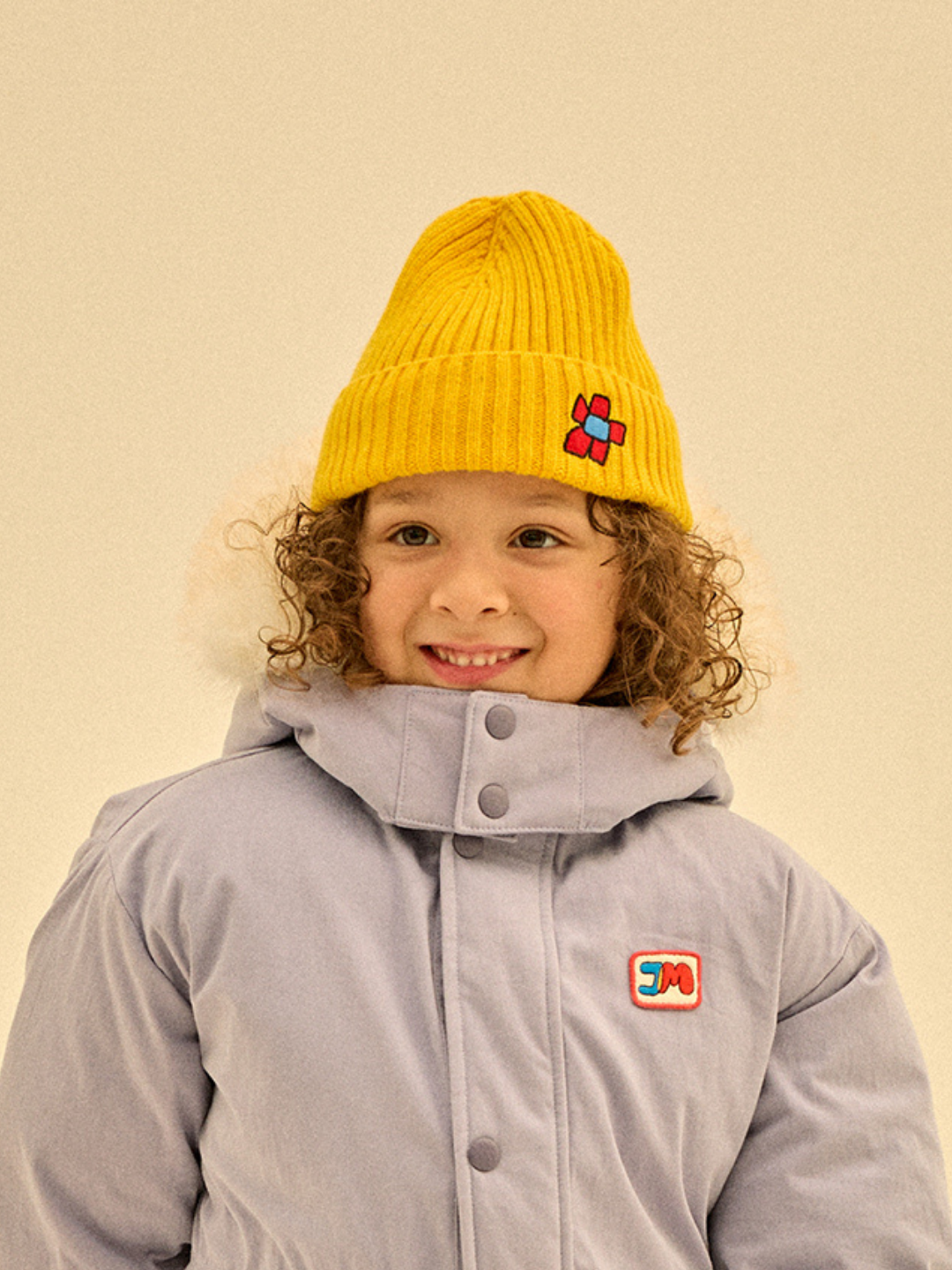 A child in a gray winter coat and a FLOWER BEANIE smiles warmly while looking off-camera, embodying the care and joy of the season.