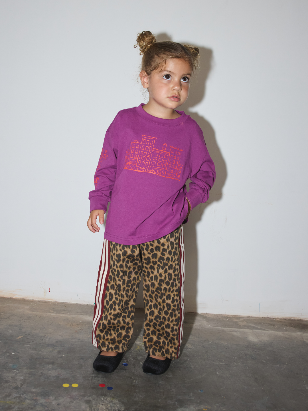 A young child standing against a plain white wall wearing a purple long-sleeve top with a pink graphic and leopard-print pants with red side stripes.