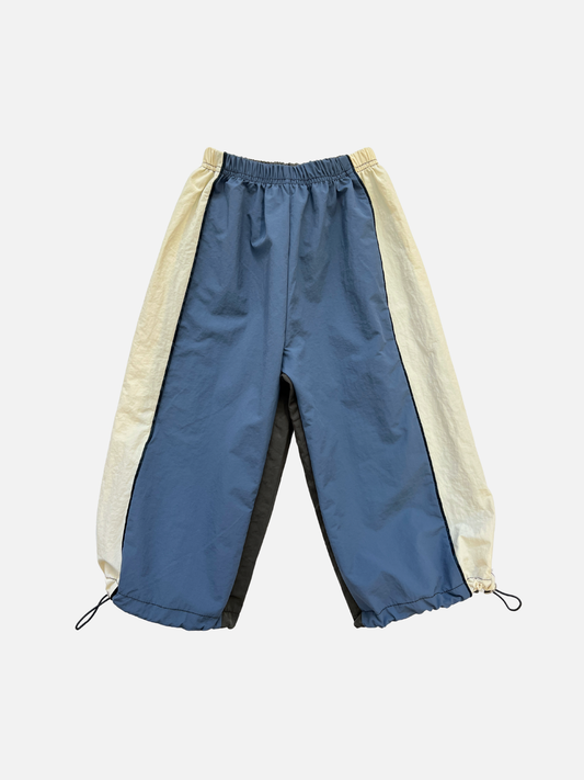 Image of The WARM-UP PANT features blue front panels, off-white sides, black back panels, and elastic waistband and cuffs—ideal for a sporty look inspired by top Korean kids brands.