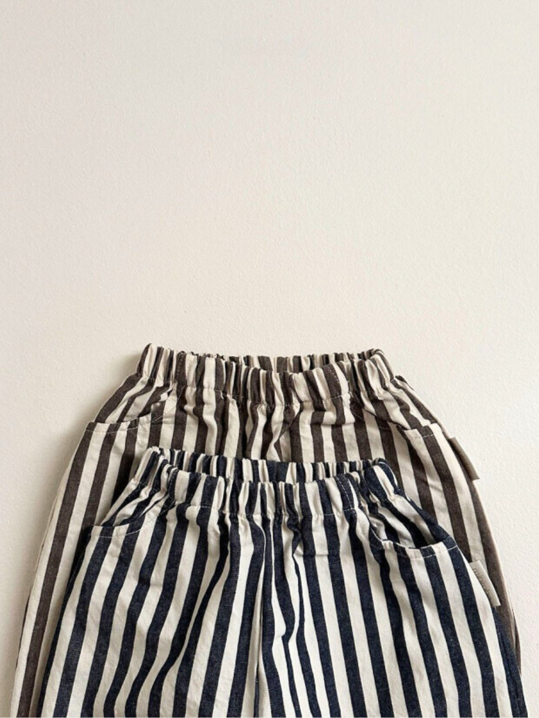 Navy Stripe | Two pairs of Minirobe's PIRATE PANTS are displayed, one featuring black and white vertical stripes and the other showcasing grey and white vertical stripes, laid flat against a plain background. Their elastic waistbands ensure comfort, while the subtle balloon leg design offers a playful touch.