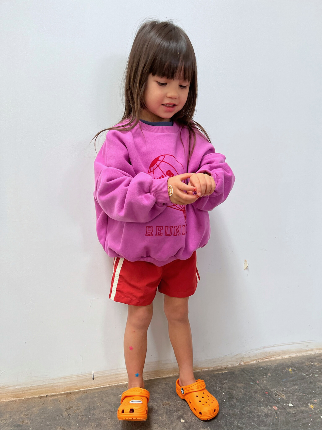 Vintage Red | A young child indoors looks down at their hands while wearing red SPRINTER SHORTS, a purple sweatshirt, and orange sandals.