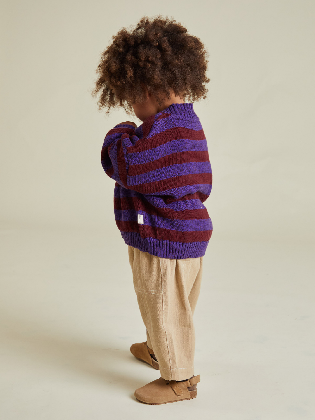 A young child with curly hair wears the EBEN PURPLE STRIPE CARDIGAN, beige pants, and brown shoes, standing sideways against a plain background—clothing cost excludes shipping and tariffs.