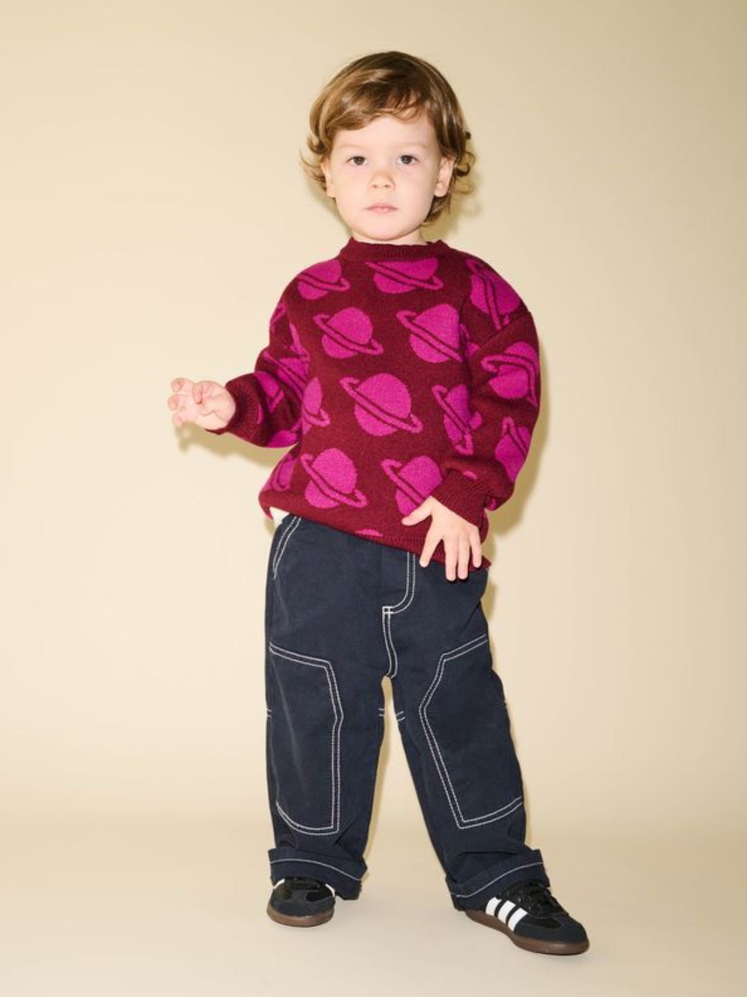 Maroon | A child with light brown hair stands against a beige background wearing the SPACE CAMP SWEATER, a maroon top adorned with planet patterns, navy pants featuring white stitching, and black sneakers with white stripes. This precise description captures the whimsical outfit in vivid detail.