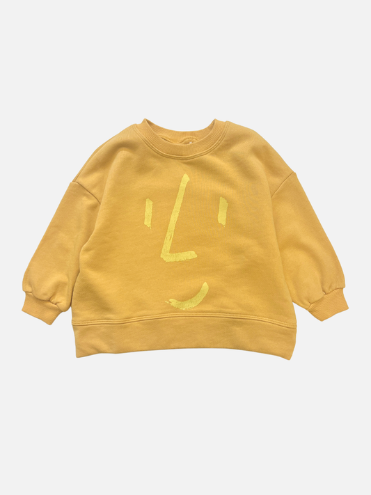 Image of The FACE SWEATSHIRT is a yellow kids' sweatshirt with a simple, abstract face design in light yellow on the front, made from sustainable materials and shown laid flat on a white background.