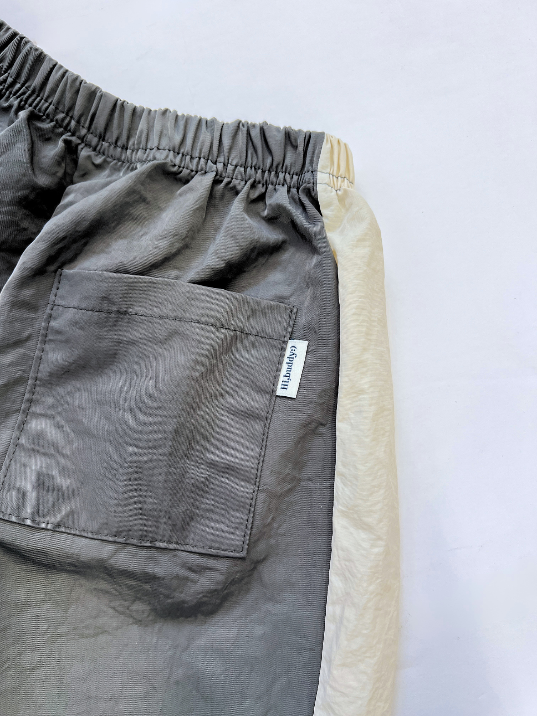 A close-up of the WARM-UP PANT—gray wide-leg track pants featuring an elastic waistband, side pocket with attached small Korean kids brand tag, and a white fabric panel along the seam.