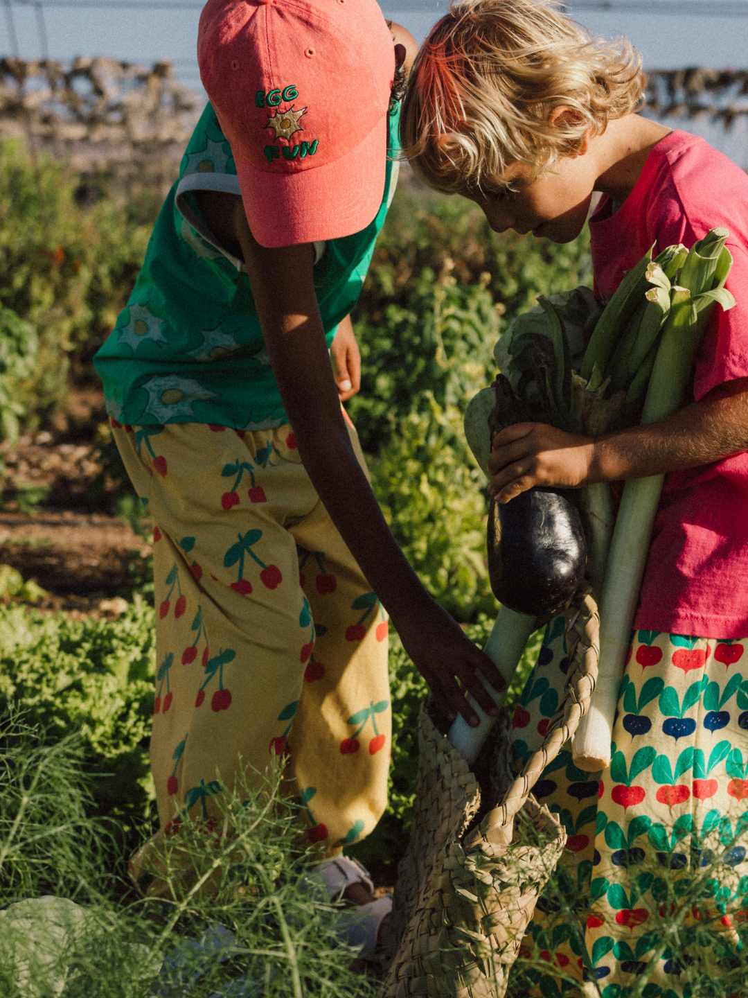 In a lush garden on a sunny day, two children wearing CHERRY PUFFY PANTS gather vegetables like leeks and an eggplant.