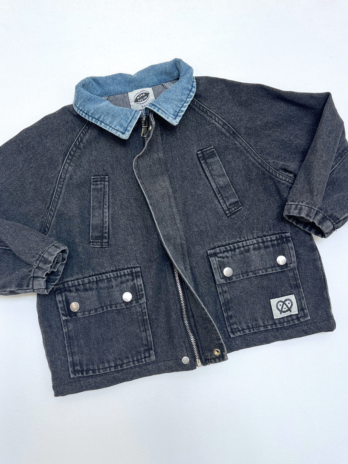 A washed black denim kids jacket with a contrasting light blue collar, laid flat on a white background. The jacket features a front zipper, vertical chest pockets, and two large front flap pockets with silver snap buttons.
