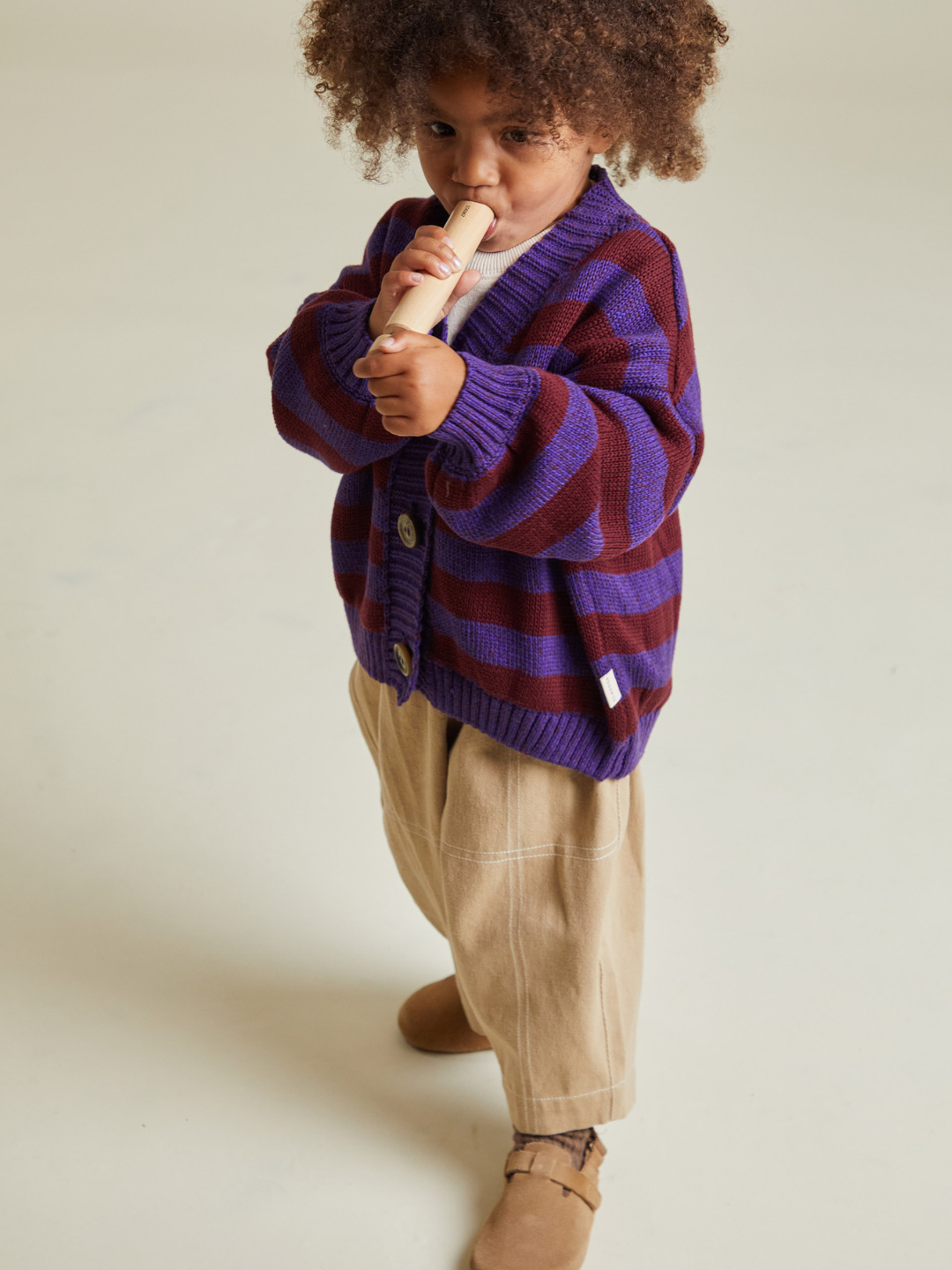A young child stands playing a wooden recorder, dressed in the EBEN PURPLE STRIPE CARDIGAN, beige pants, and brown shoes—a charming moment showing that joy is truly priceless.