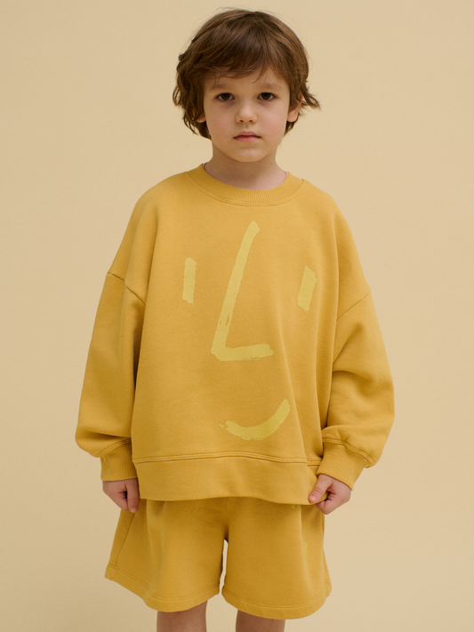 Second image of The FACE SWEATSHIRT is a yellow kids' sweatshirt with a simple, abstract face design in light yellow on the front, made from sustainable materials and shown laid flat on a white background.