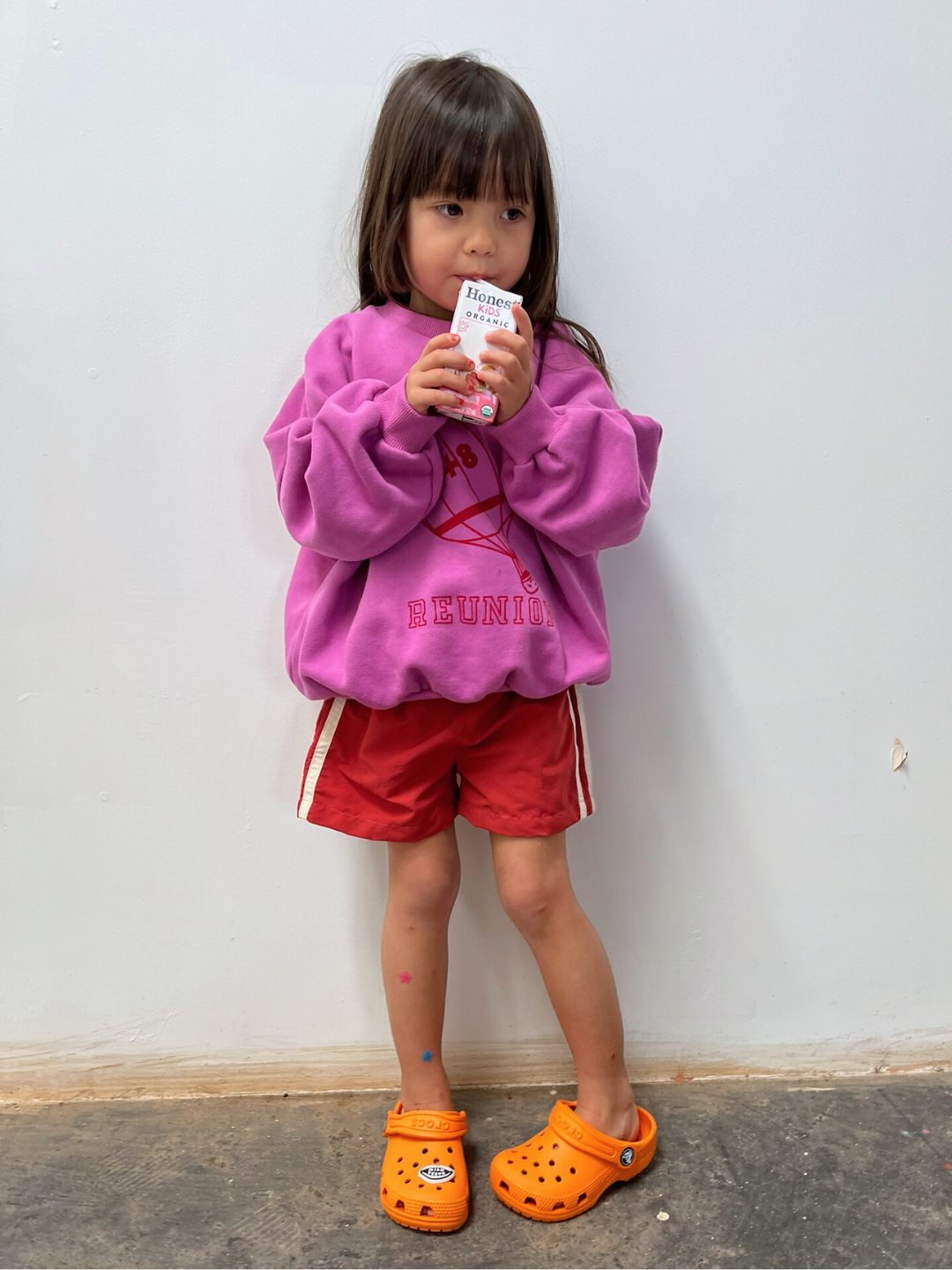 A young child in a pink sweatshirt, SPRINTER SHORTS, and orange Crocs stands against a plain wall, sipping from a juice box.