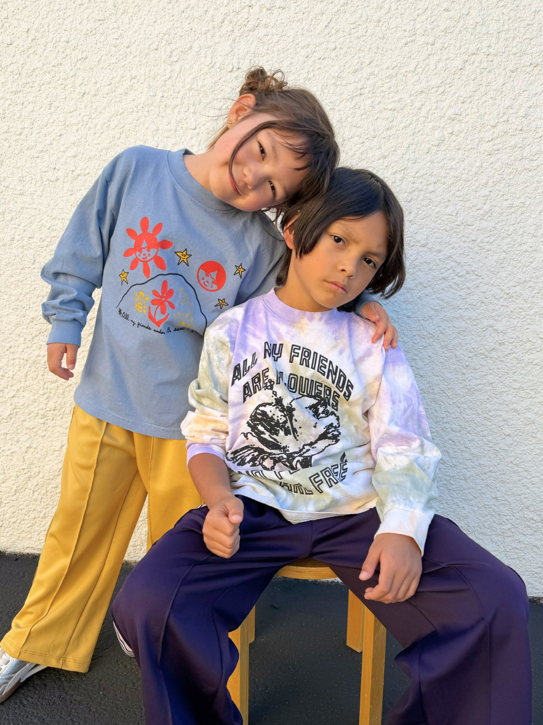Multi | Two children posing against a white wall, one sitting on a chair and the other standing.
