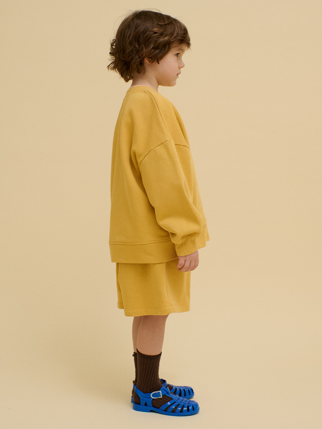 A young child stands in profile against a beige background, wearing the sustainable FACE SWEATSHIRT in mustard yellow with matching shorts, brown socks, and blue sandals.