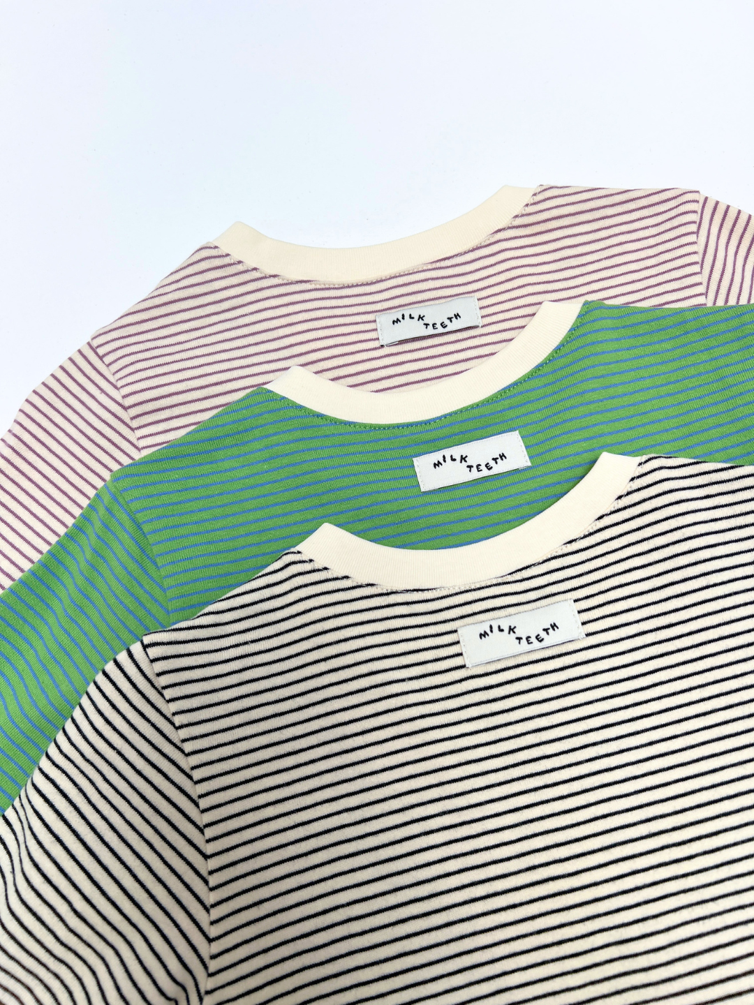 Three BABY STRIPE LONGSLEEVE shirts in purple, green, and black are arranged overlapping on a white surface, each with a "Nile Teeth" label at the neckline. Made from soft cotton jersey for easy dressing, these baby and toddler basics blend comfort with playful style.