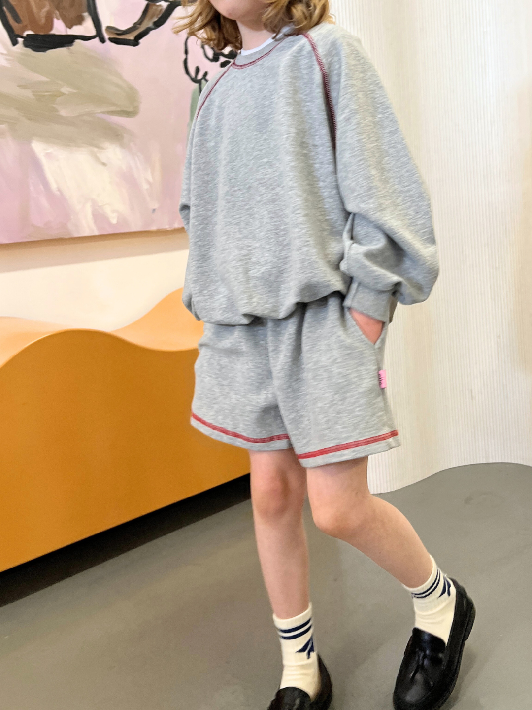 A child in a gray sweatshirt and VARSITY SWEAT SHORTS featuring vintage-inspired brushed cotton with red trim, black shoes, and white socks stands indoors before abstract art and a wooden bench, embodying a timeless Korean kids brand style.