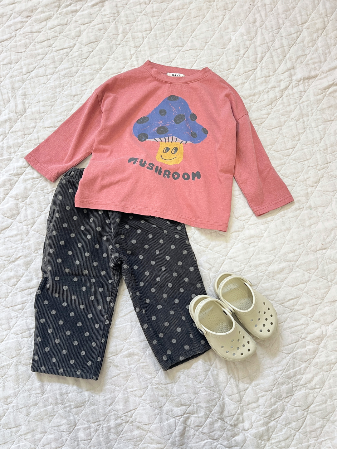 Children's outfit with a MUSHROOM LONG SLEEVE TEE in pink, adorned with a blue mushroom graphic, paired with gray polka dot pants and white slip-on shoes, all set on a white quilted background.