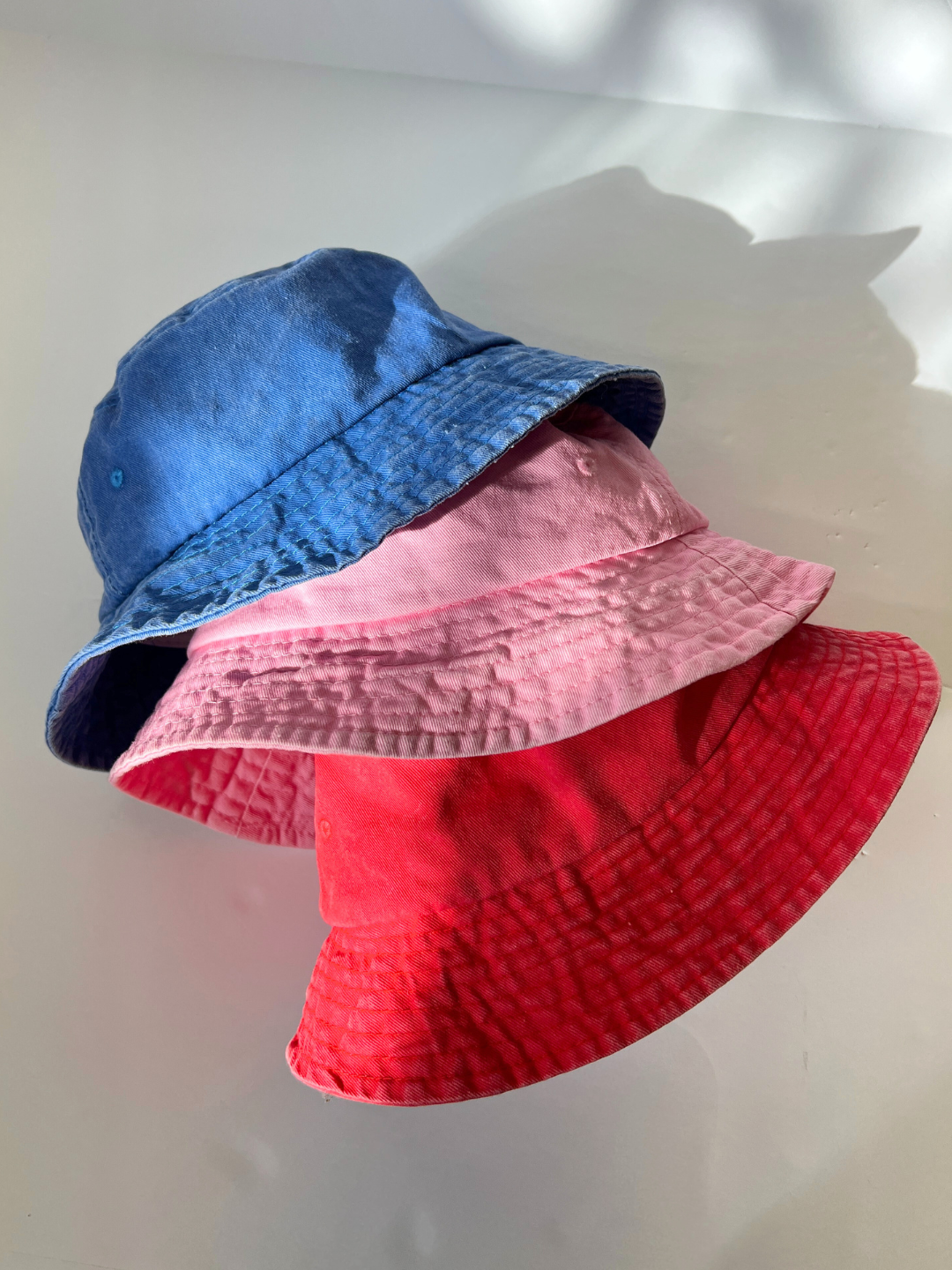 Red | Three PIGMENT BUCKET HATs, exuding vintage charm from their dye, are elegantly arranged on a clean white surface—blue on top, pink in the middle, and bold red at the base.