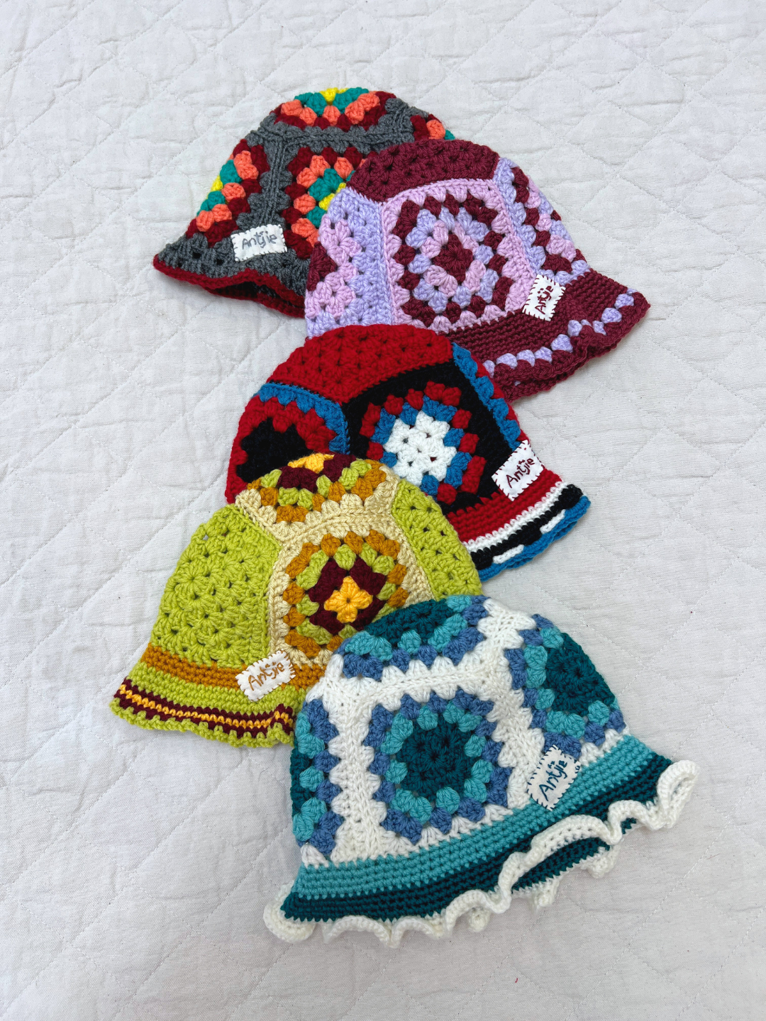 Four vibrant CROCHETED BUCKET HAT 1-3Y pieces with granny square patterns are displayed in a row on a white quilted surface, reflecting colorful South Africa-inspired craftsmanship.