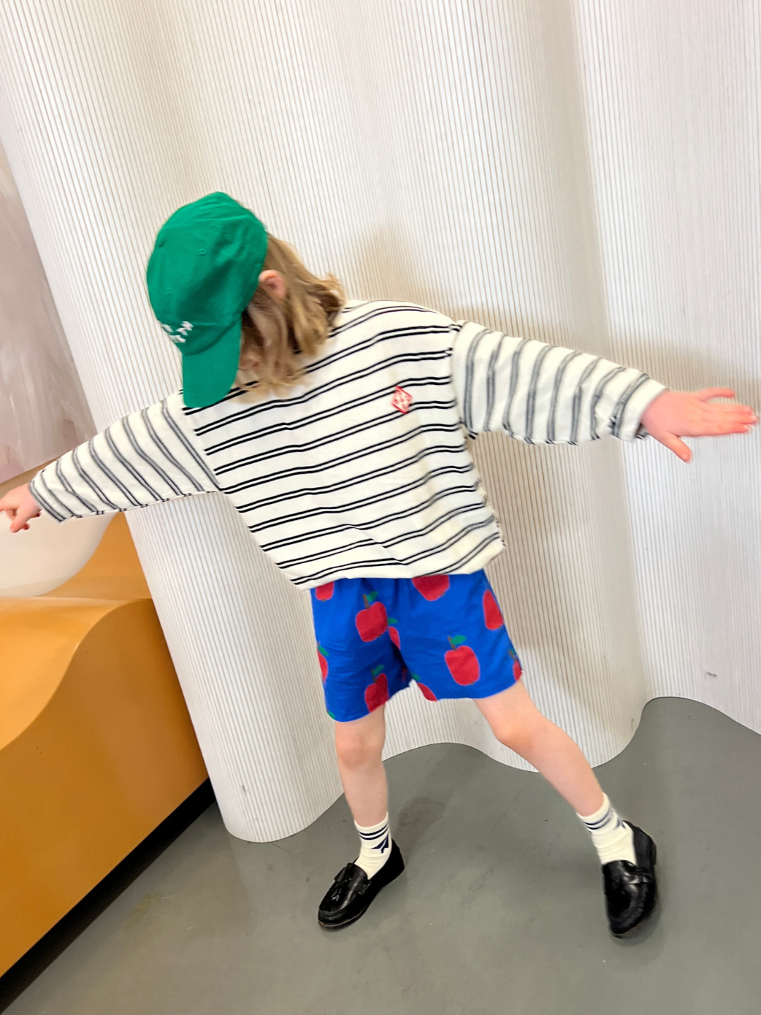 A child in a green cap and the PEPPER LONGSLEEVE TEE from a Korean kids brand, wearing blue apple print shorts, white socks, and black shoes, stands with arms outstretched in front of a curved wall and yellow seat.