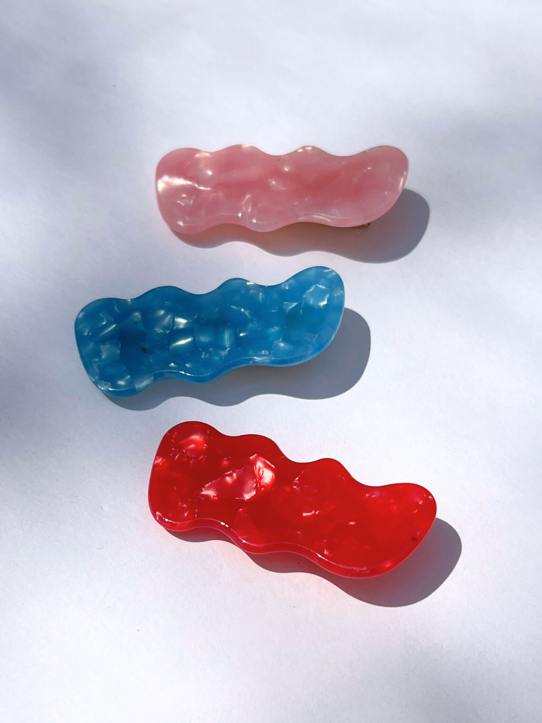 The SQUIGGLE HAIRCLIP SET features three wavy, rectangular resin barrettes in pink, blue, and red, arranged vertically on a white background as vibrant hair accessories.