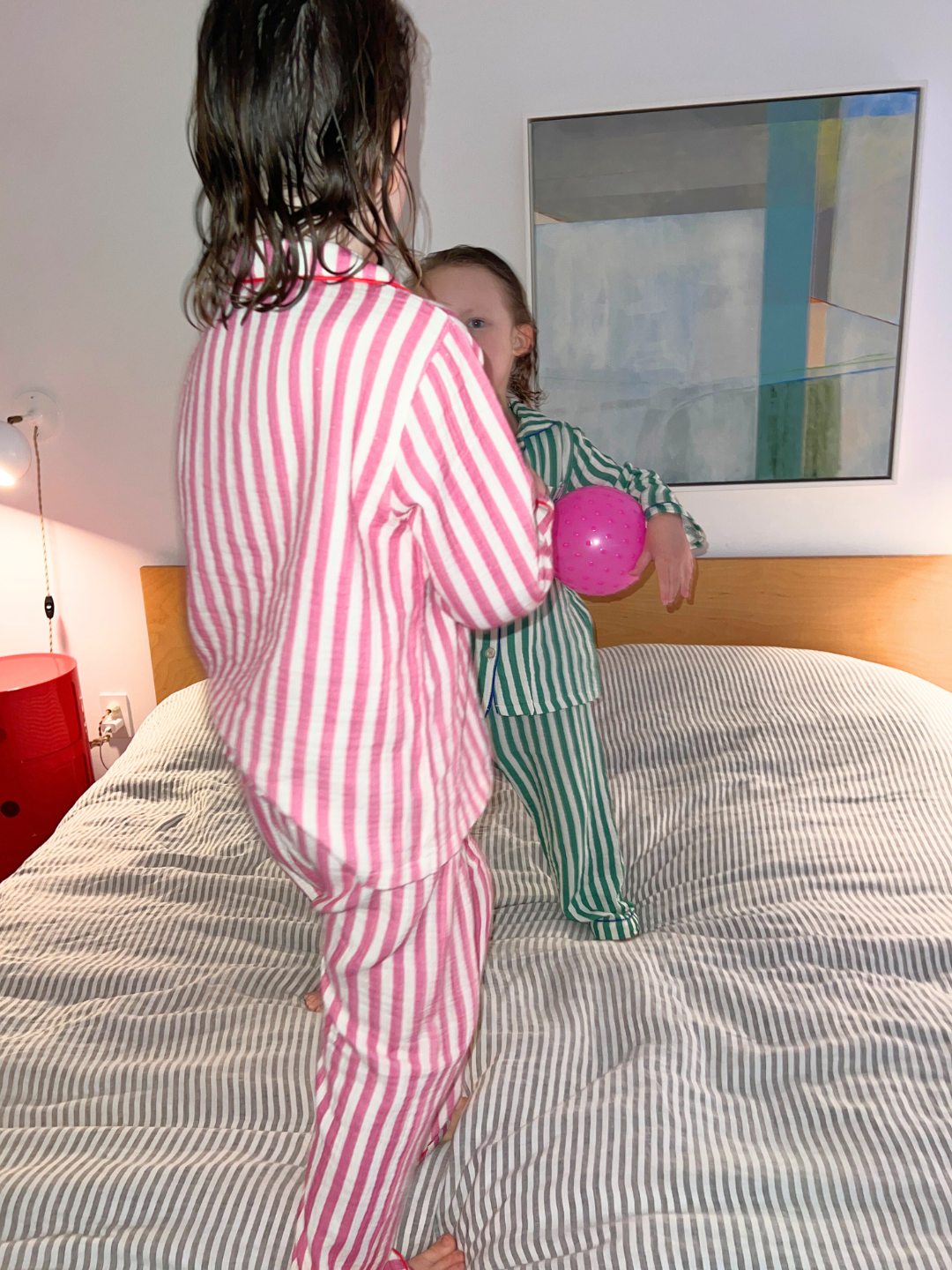 Two children in the MOVIE NIGHT SET, a striped cotton muslin ensemble, are on a bed. One holds a pink ball. The room is cozy with a painting on the wall and a red nightstand.