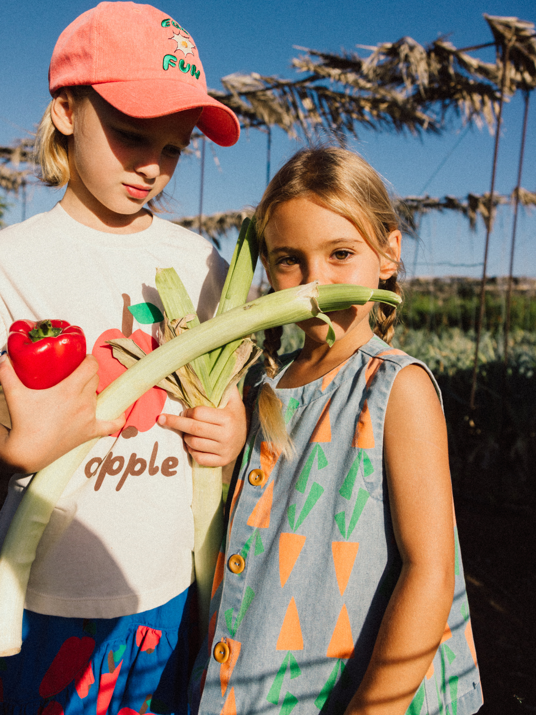 Two children stand in a sunny garden. One holds a red pepper and a leek, while the other wears the CARROT DENIM VEST. A framework of dried plants is visible in the background.