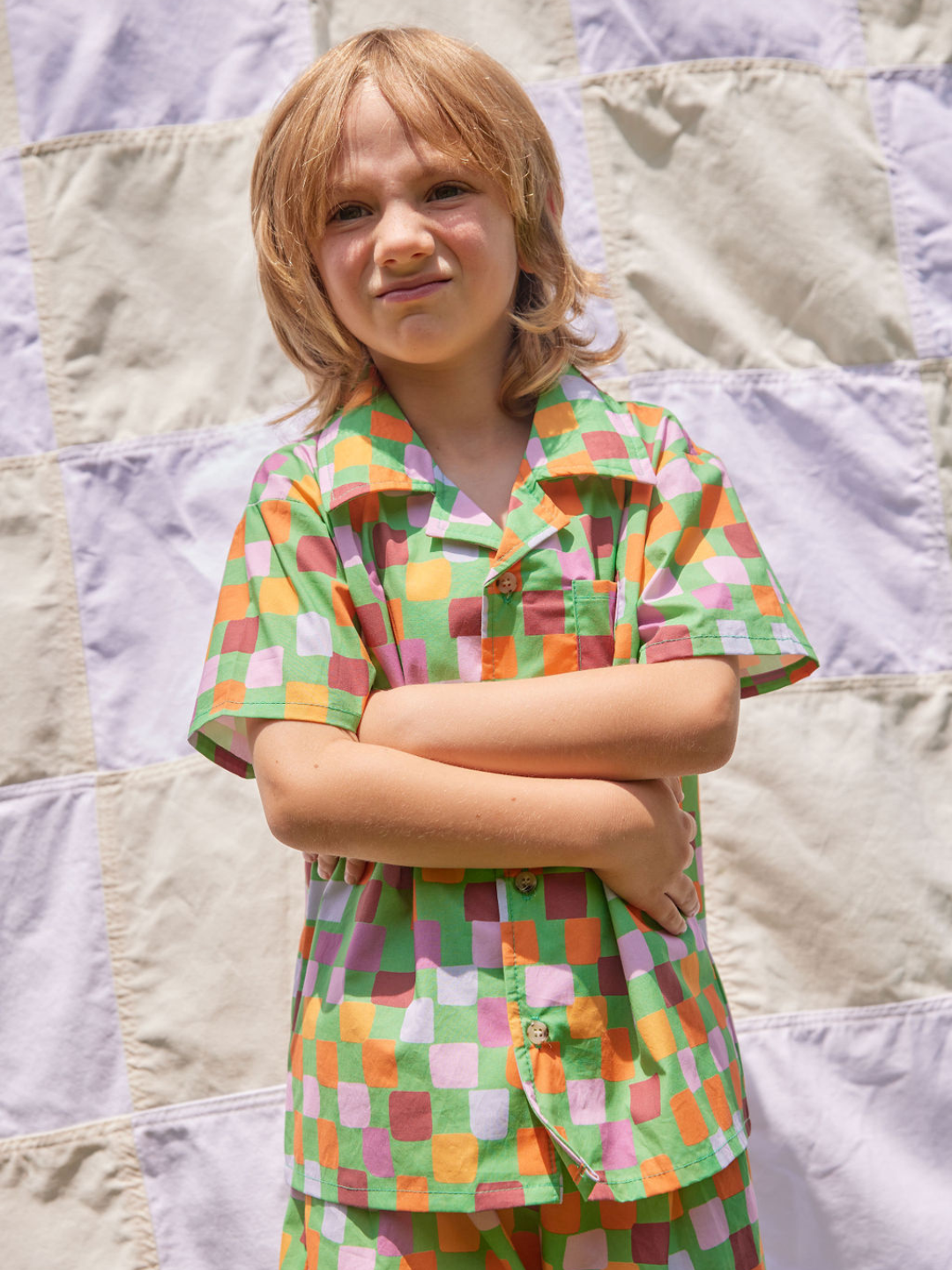 A child wearing a kids' shirt and shorts set in a pattern of purple, pink, gold and orange squares on a green background